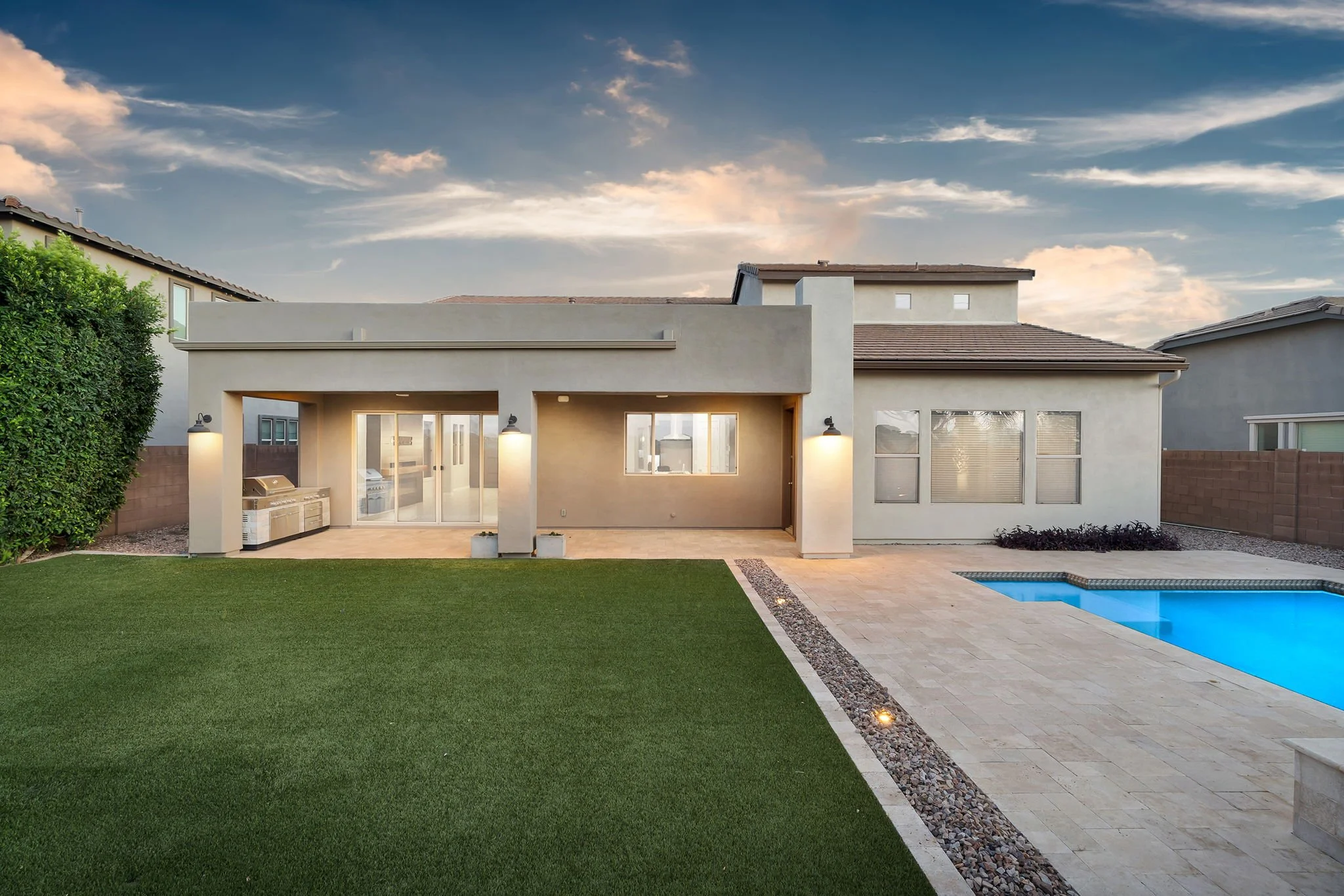 Modern white house with backyard patio, swimming pool, landscaped lawn, and cloudy sky at dusk.