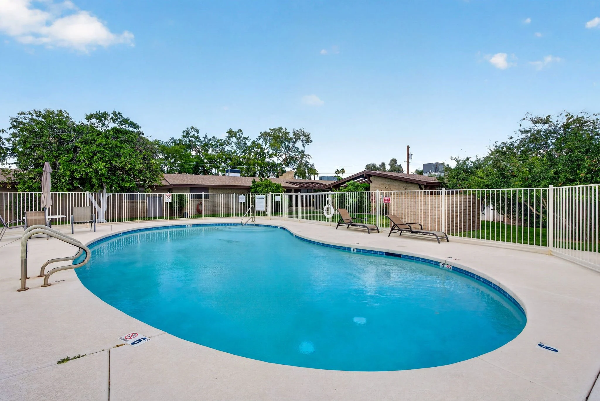 An outdoor swimming pool with a concrete deck, surrounded by a white fence, with lounge chairs and an umbrella. There are trees and buildings in the background under a blue sky with a few clouds.