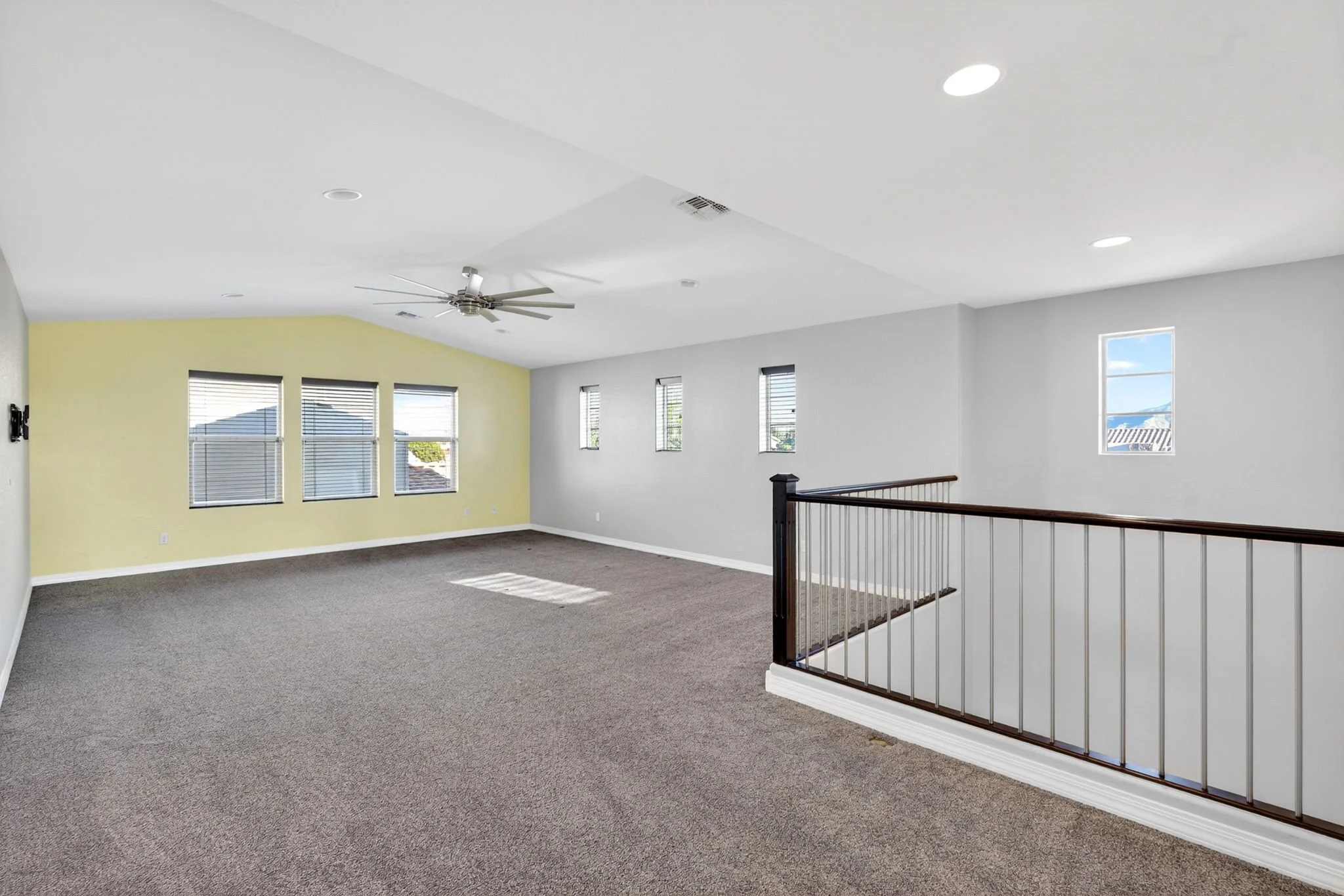 An open, spacious room with beige carpet and a white ceiling with recessed lighting and a ceiling fan. The walls are painted yellow and light gray, with multiple windows letting in natural light. There is a staircase with a black metal and wood raili