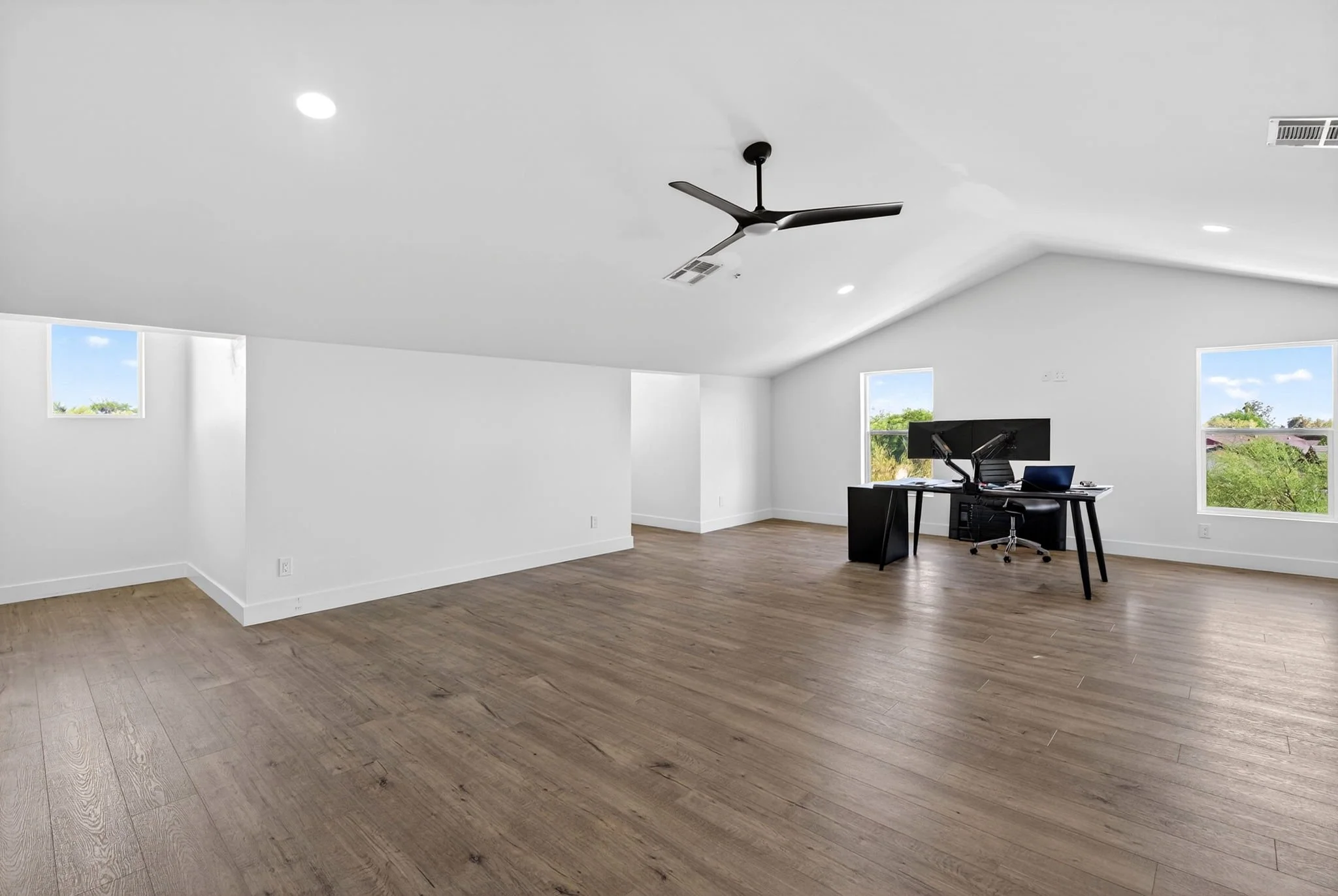Empty modern room with large windows, white walls, wooden floor, ceiling fan, and a desk with computer.