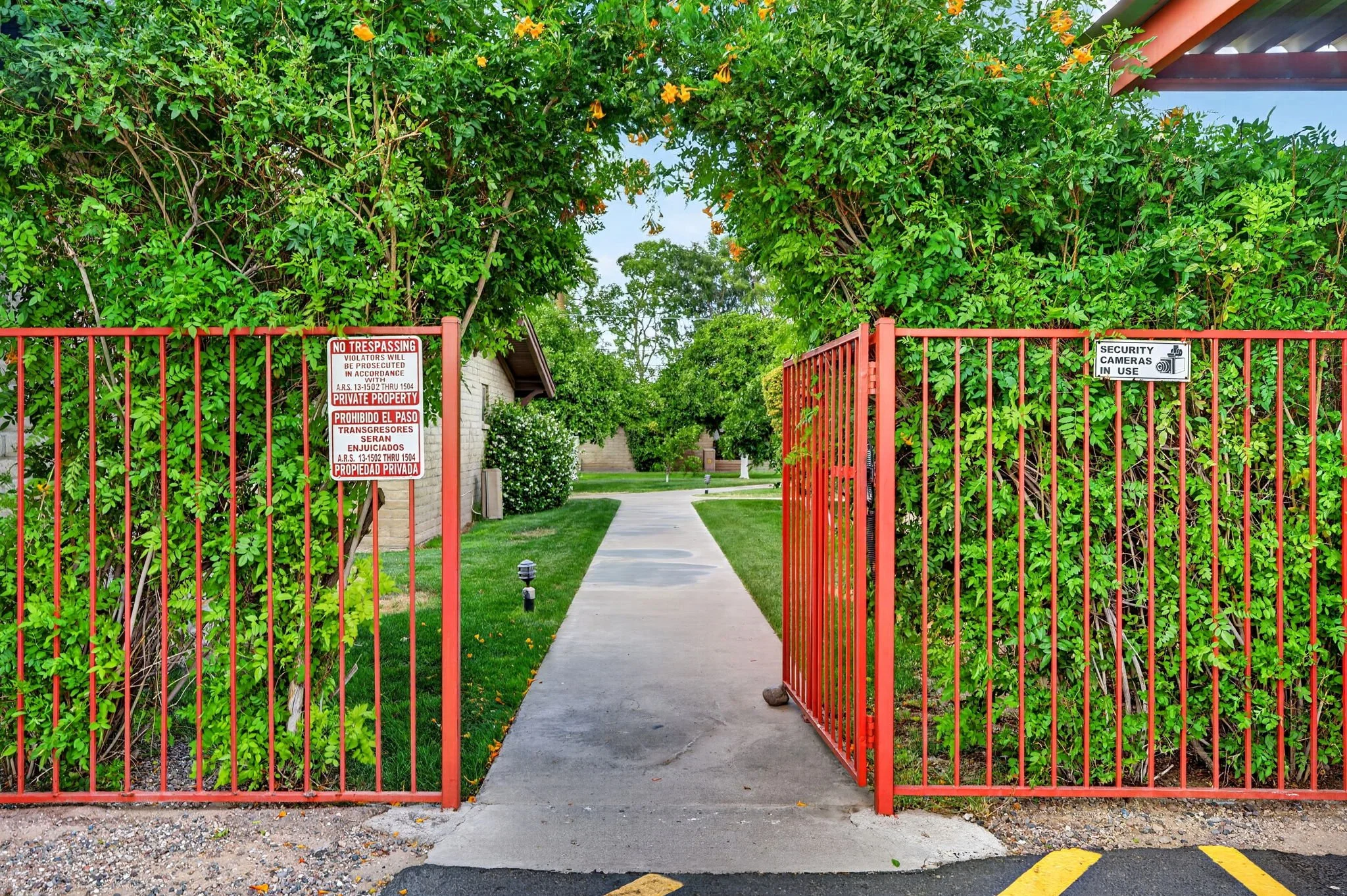 Open red gate leading to a sidewalk and grassy area surrounded by tall green leafy bushes with yellow flowers. Signs on the gate indicate no trespassing and security cameras in use.