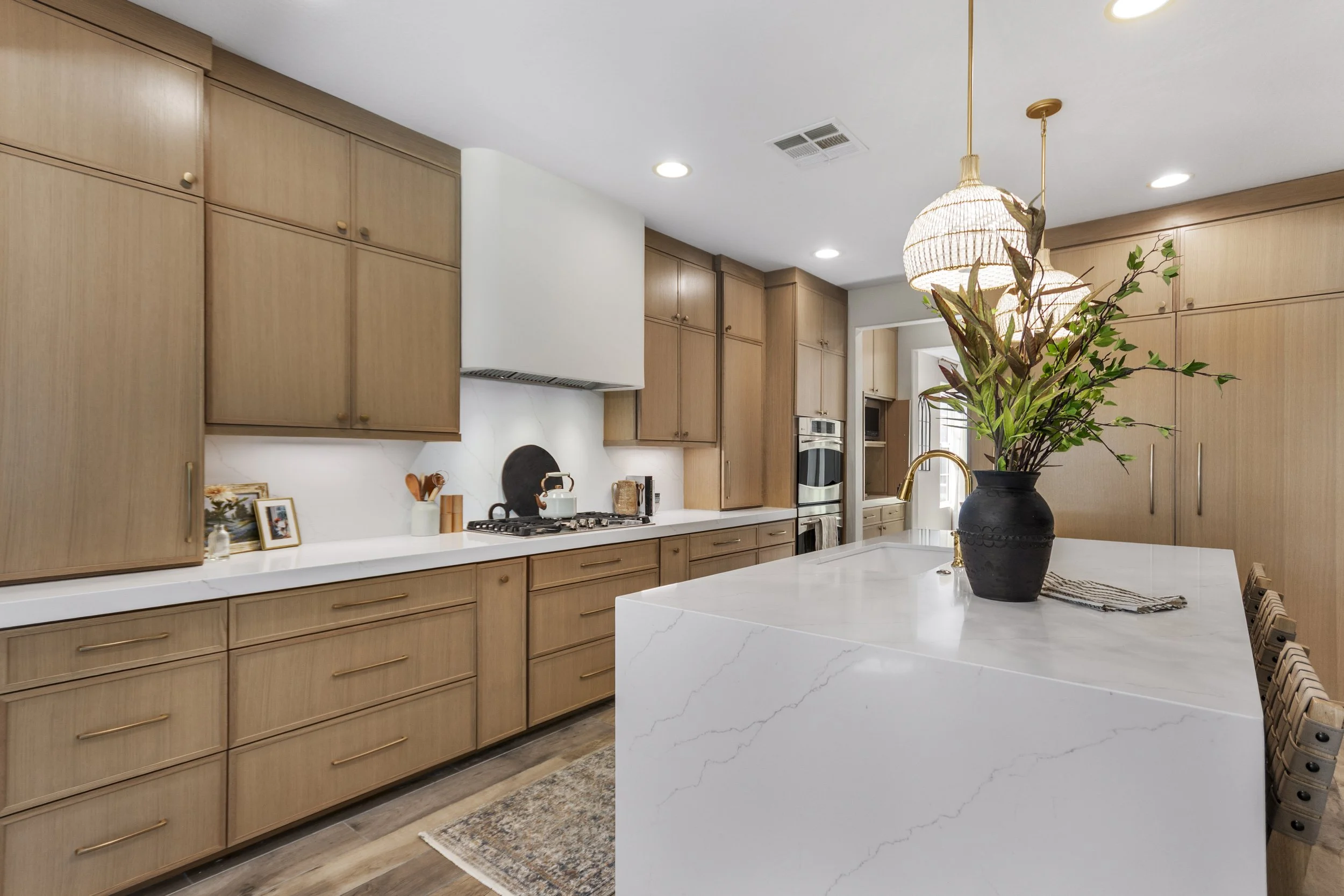 Contemporary kitchen with light wood cabinets, white marble countertops, and a kitchen island with a black vase and green plant. Pendant light overhead and built-in stainless steel oven visible in the background.