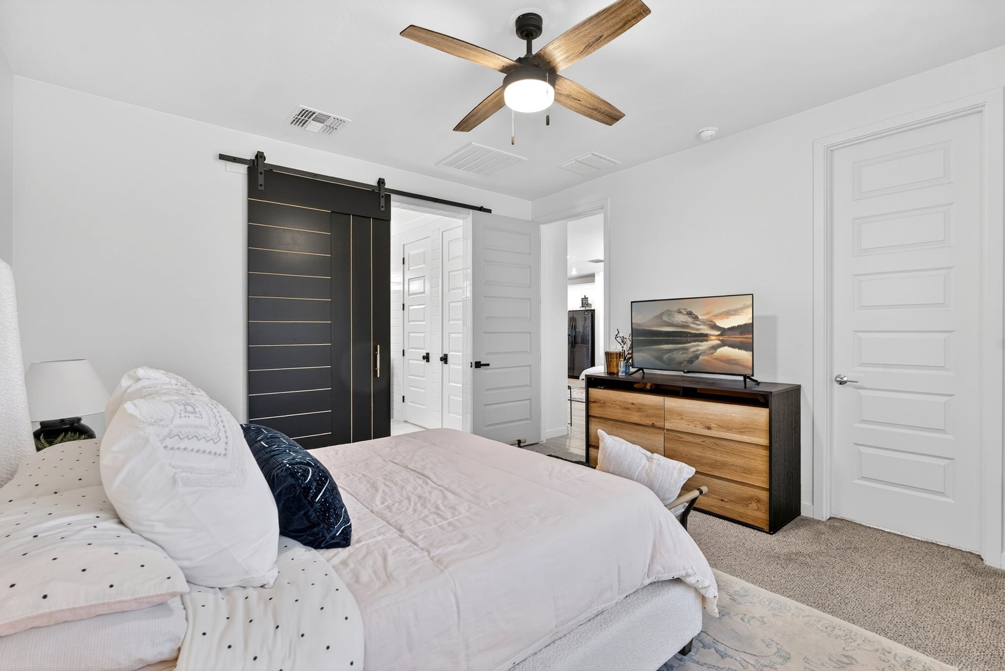 A bedroom with white walls, a bed with white polka dot bedding and pillows, a black sliding barn door, a wooden dresser with a flat-screen TV, and a ceiling fan with wooden blades.