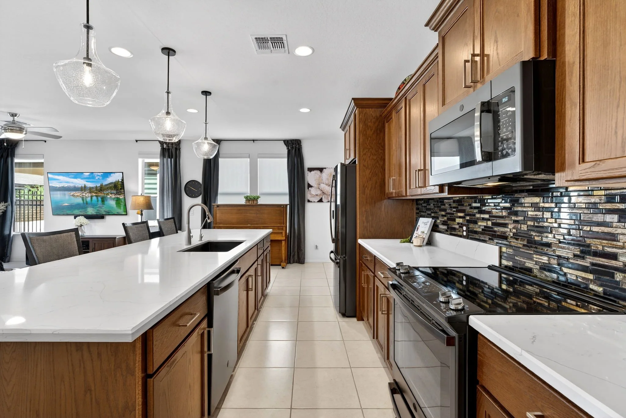 A modern kitchen with wooden cabinets, a white island with a sink, stainless steel appliances, a black stove, a microwave, and a backsplash with black and metallic tiles. The room has light-colored floor tiles, and there are large windows with dark c