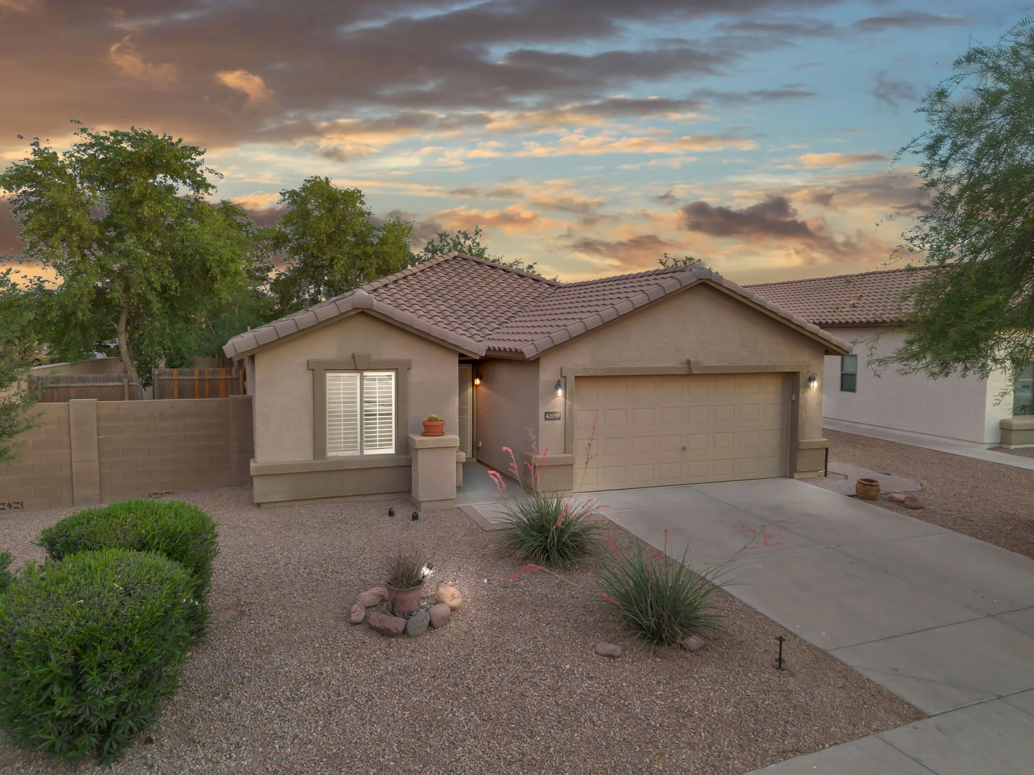 A single-story house with a beige exterior, a tiled roof, and a garage door. There is a small patio with a potted plant and a lit wall-mounted light next to the door. The front yard has desert landscaping with dry gravel, bushes, and small rocks, and