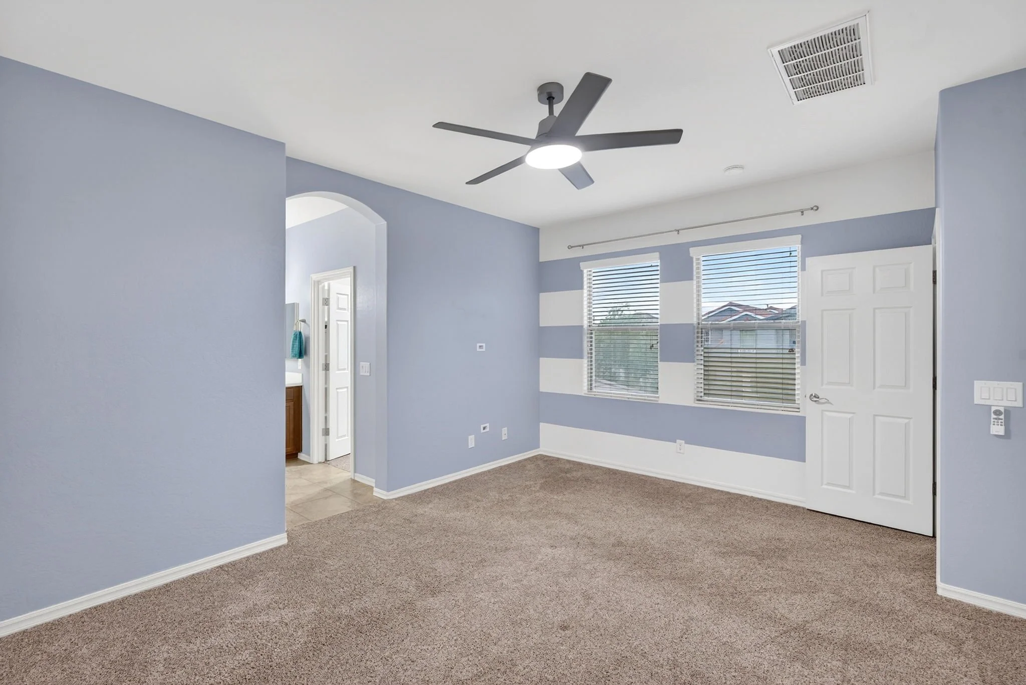 Empty living room with blue walls, large windows with blinds, ceiling fan with light, carpeted floor, and an open door leading to another room.