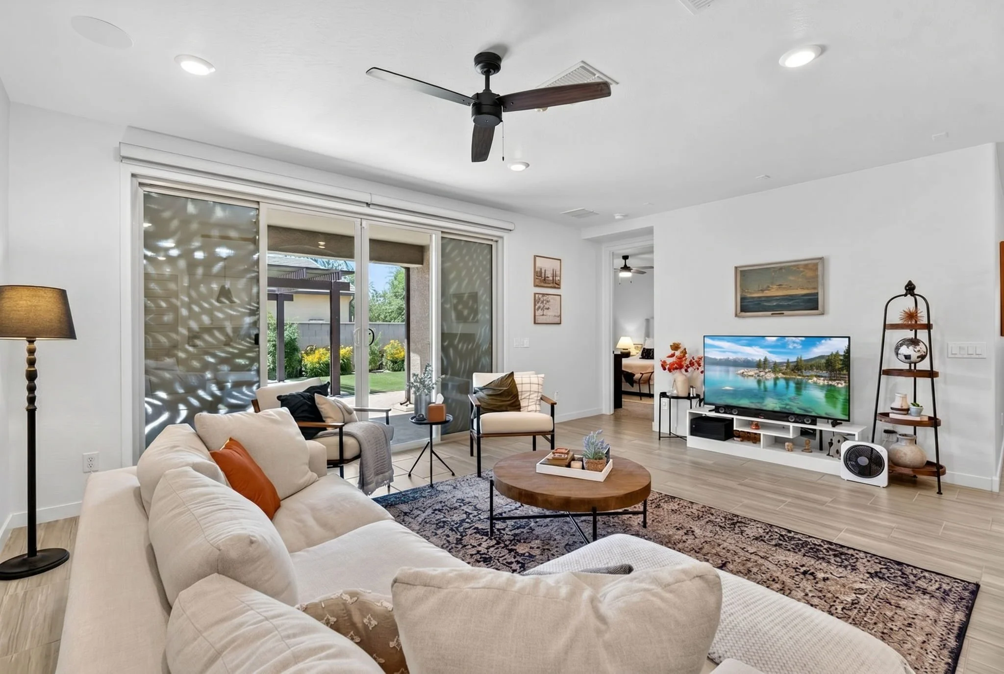 A modern living room with a large sectional sofa, a patterned rug, a wall-mounted flat-screen TV, and decorative items. There's a large glass sliding door leading to a backyard with greenery, and a ceiling fan with dark wood blades.