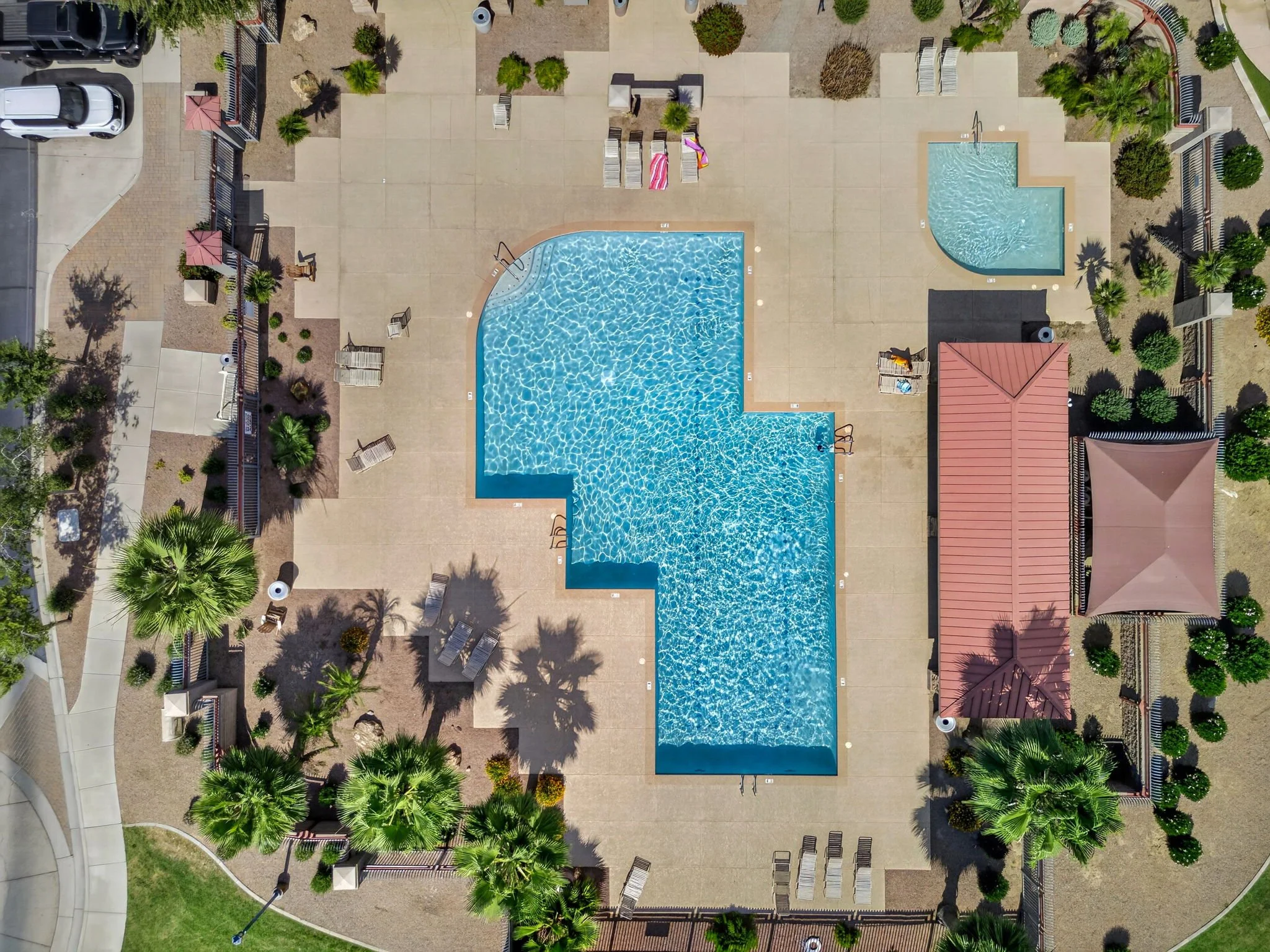 An aerial view of a community swimming pool area with surrounding lounge chairs, umbrellas, trees, and plants, enclosed by fencing and adjacent to residential structures and parked cars.