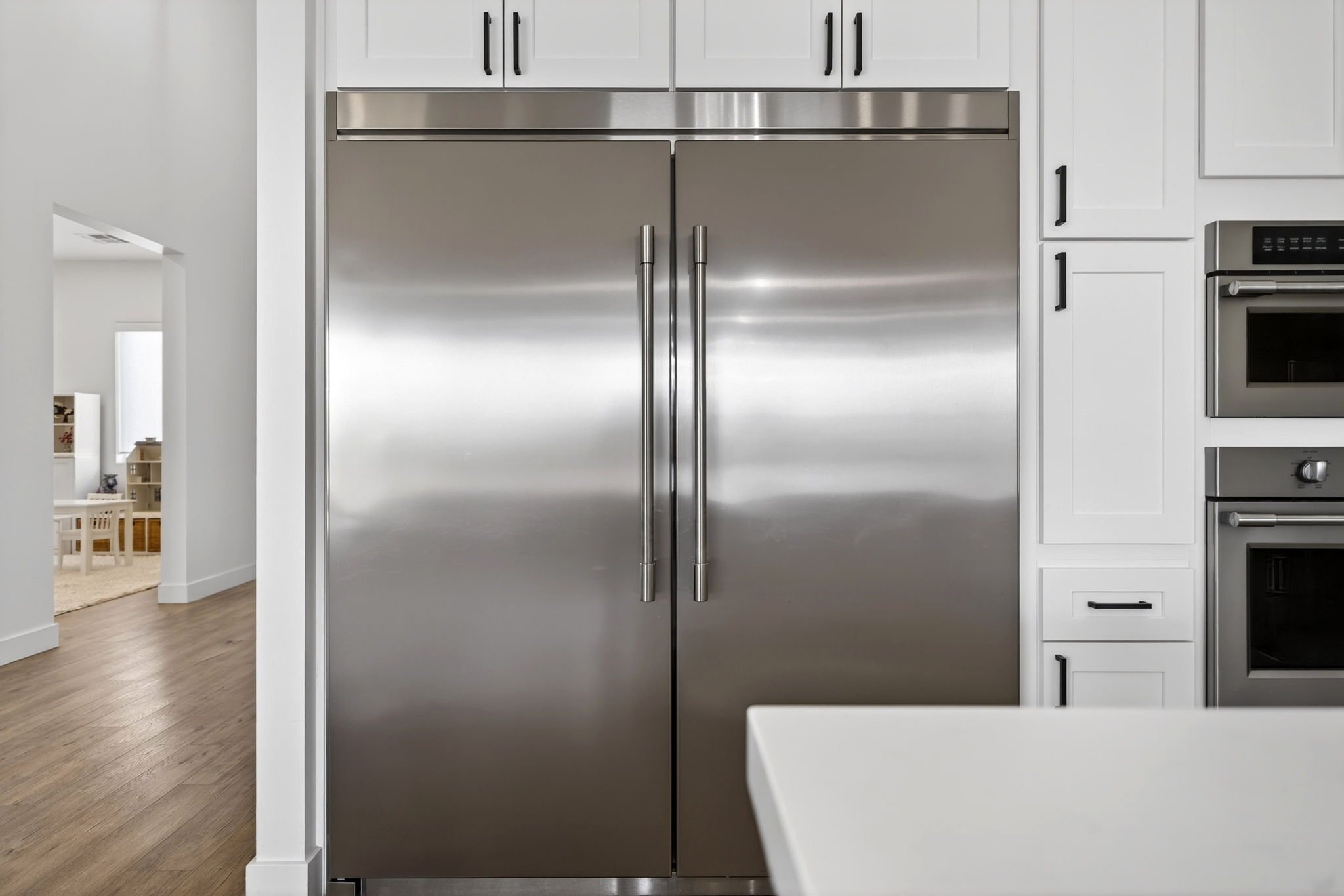 Stainless steel double-door refrigerator in a modern kitchen with white cabinets and black handles, hardwood floor, and a view into a bright dining area.