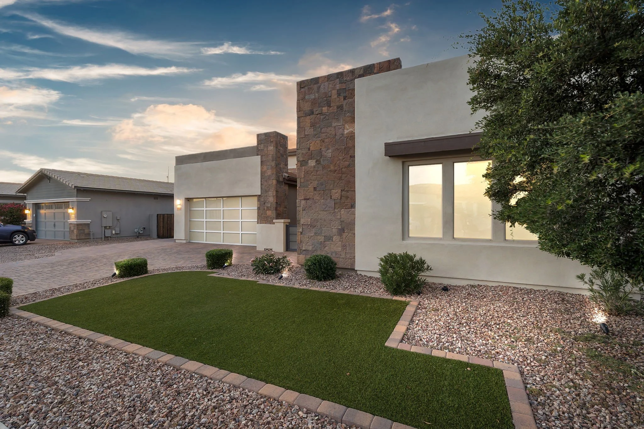 A modern residential house featuring a combination of stone and stucco exterior walls, a large window with a browned awning, a garage with a white door, a paved driveway, landscaped yard with green grass, small bushes, and decorative rocks, with a cl