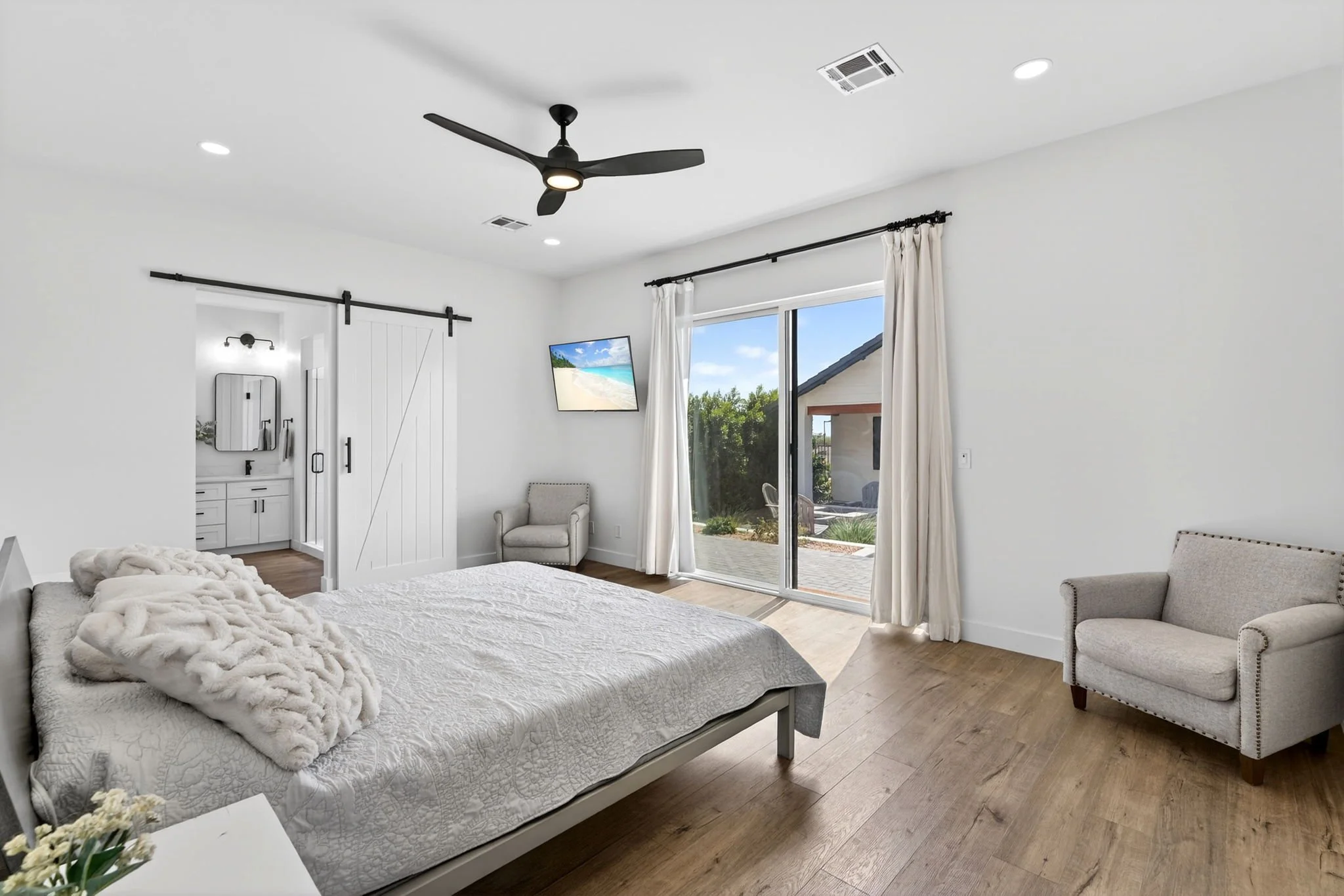 Modern bedroom with white walls, a bed with a white bedspread, a faux fur blanket, and a white bedside table with a flower arrangement. There are armchairs near a large sliding glass door with white curtains, and a wall-mounted TV displaying a beach 
