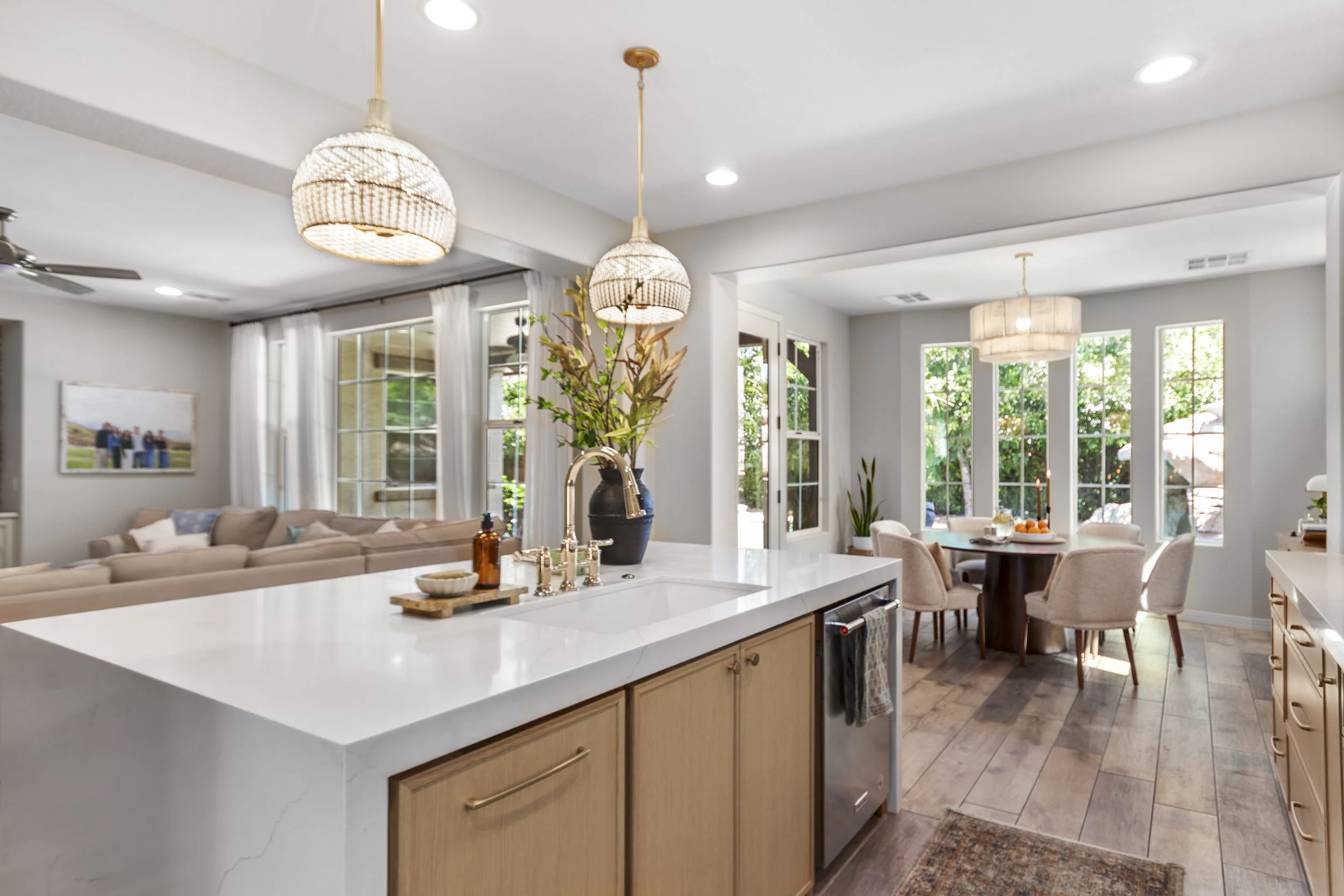 Open-concept living room and kitchen with white walls, large windows, and natural light, featuring pendant lights, a beige sofa, a wooden dining table with chairs, and green potted plants.