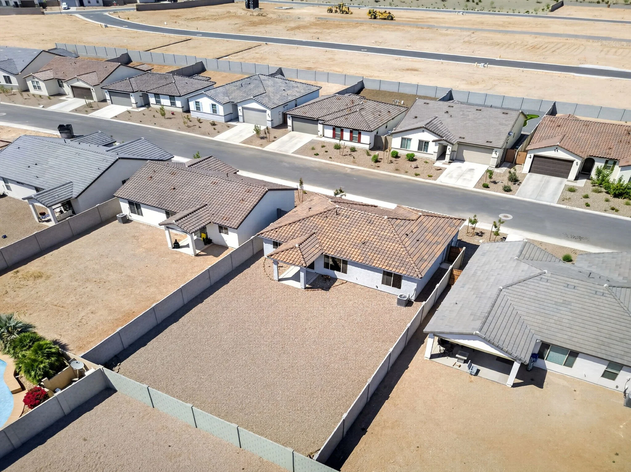 Aerial view of a suburban neighborhood with single-story houses, yellow construction equipment in the background, and a road with a sidewalk in the foreground.