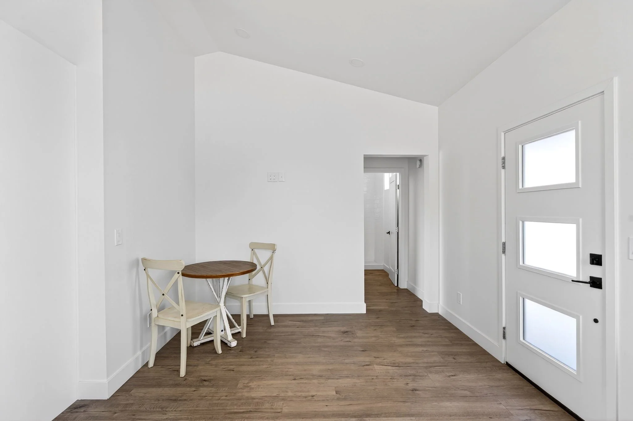 Inside view of a bright white room with a small round wooden table and two chairs, near a white door with glass panels, hardwood flooring, and small windows.