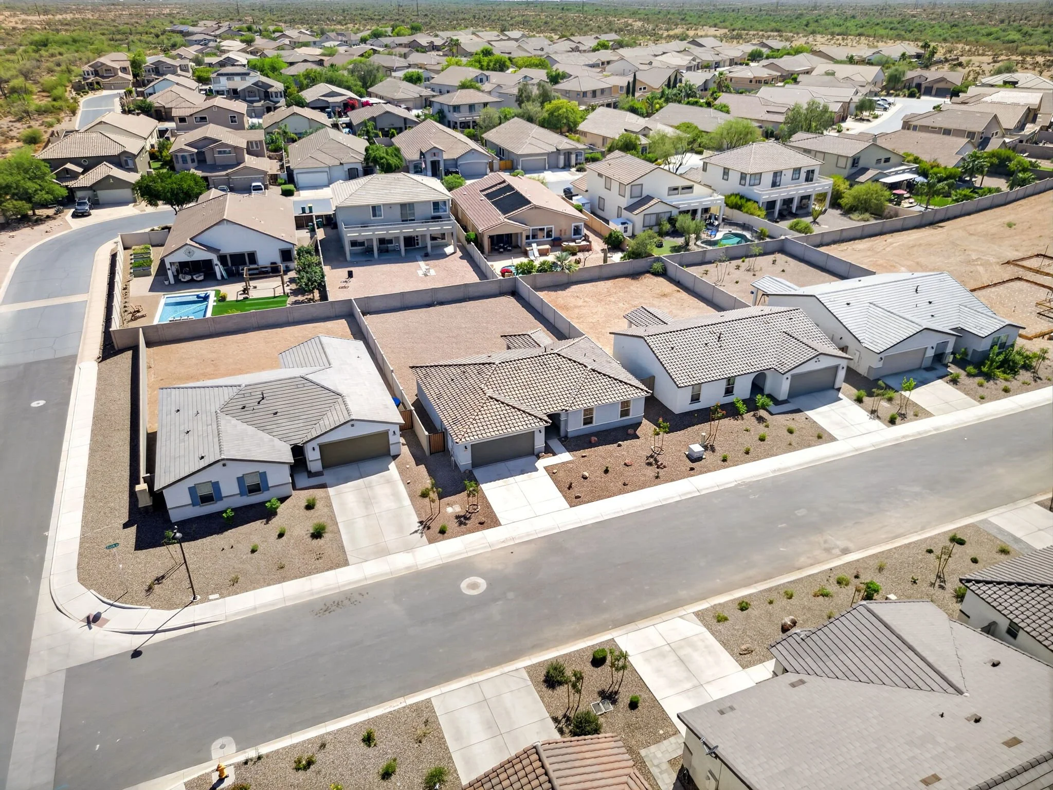 Aerial view of suburban houses with tiled roofs, some with swimming pools and landscaped yards, in a desert-like area with sparse vegetation.