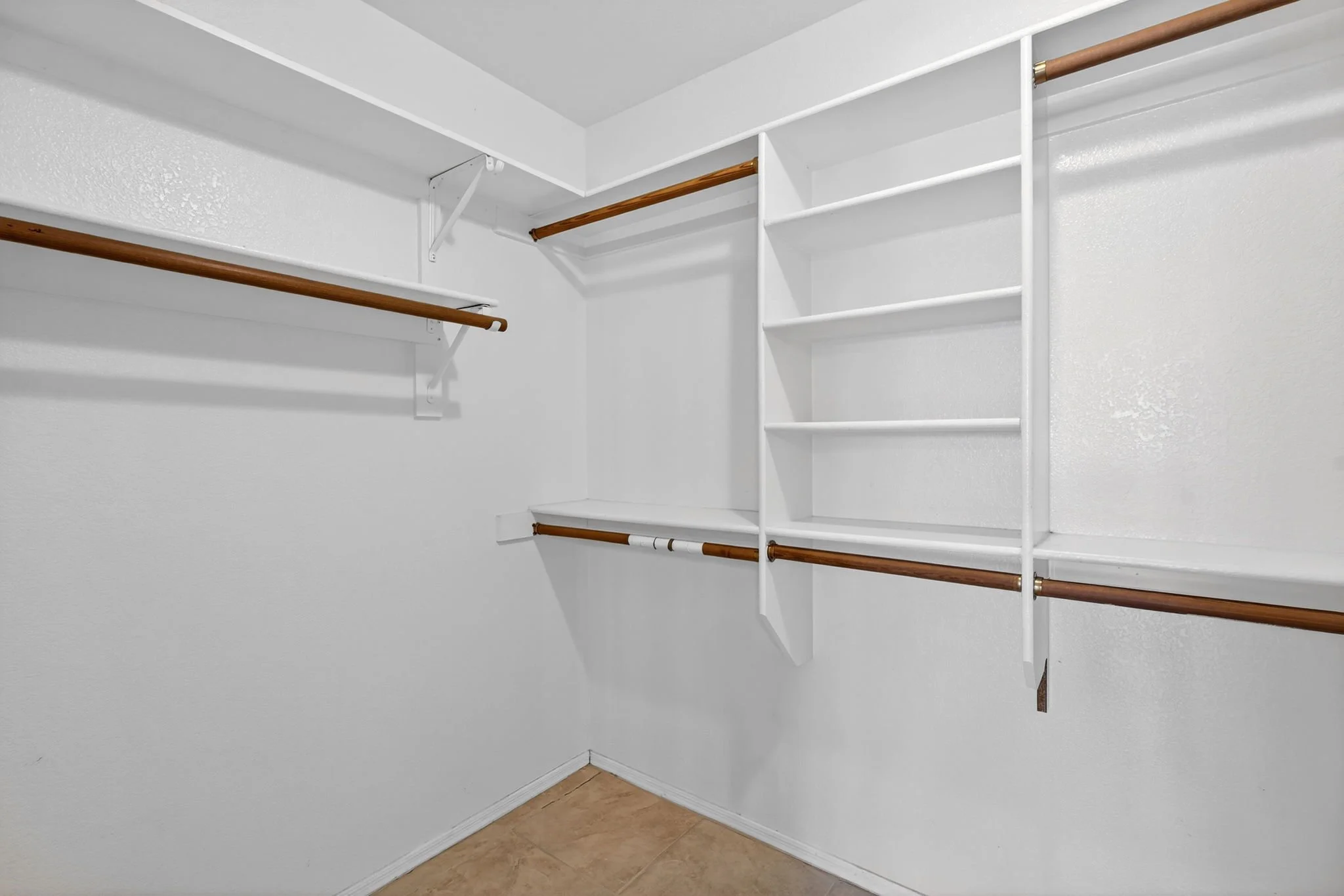 Empty walk-in closet with white built-in shelves and hanging rods, beige tile floor, and white textured walls.