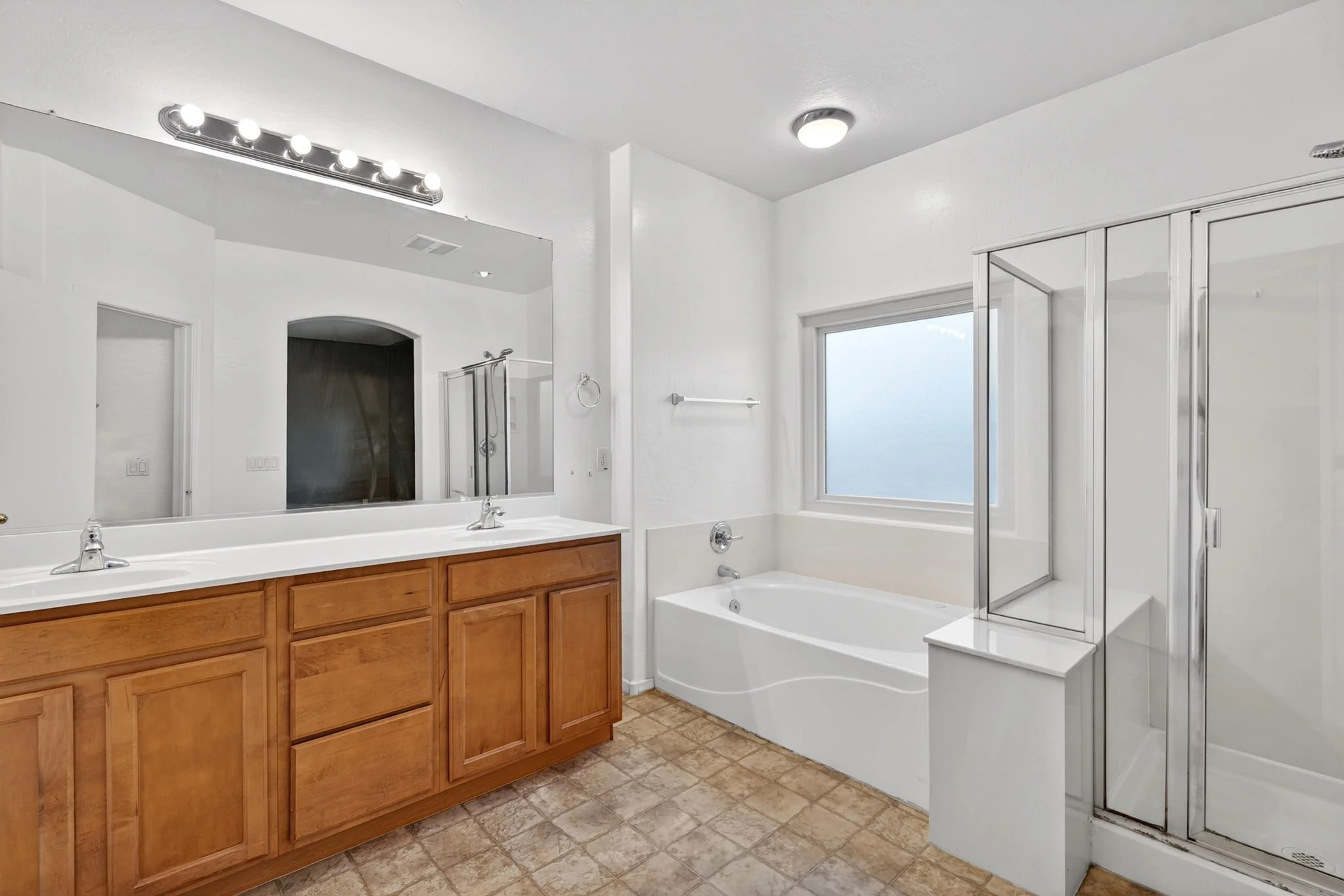 A bathroom with a large mirror, double sinks, a bathtub, a glass shower enclosure, and a window letting in natural light, with white walls and beige floor tiles.