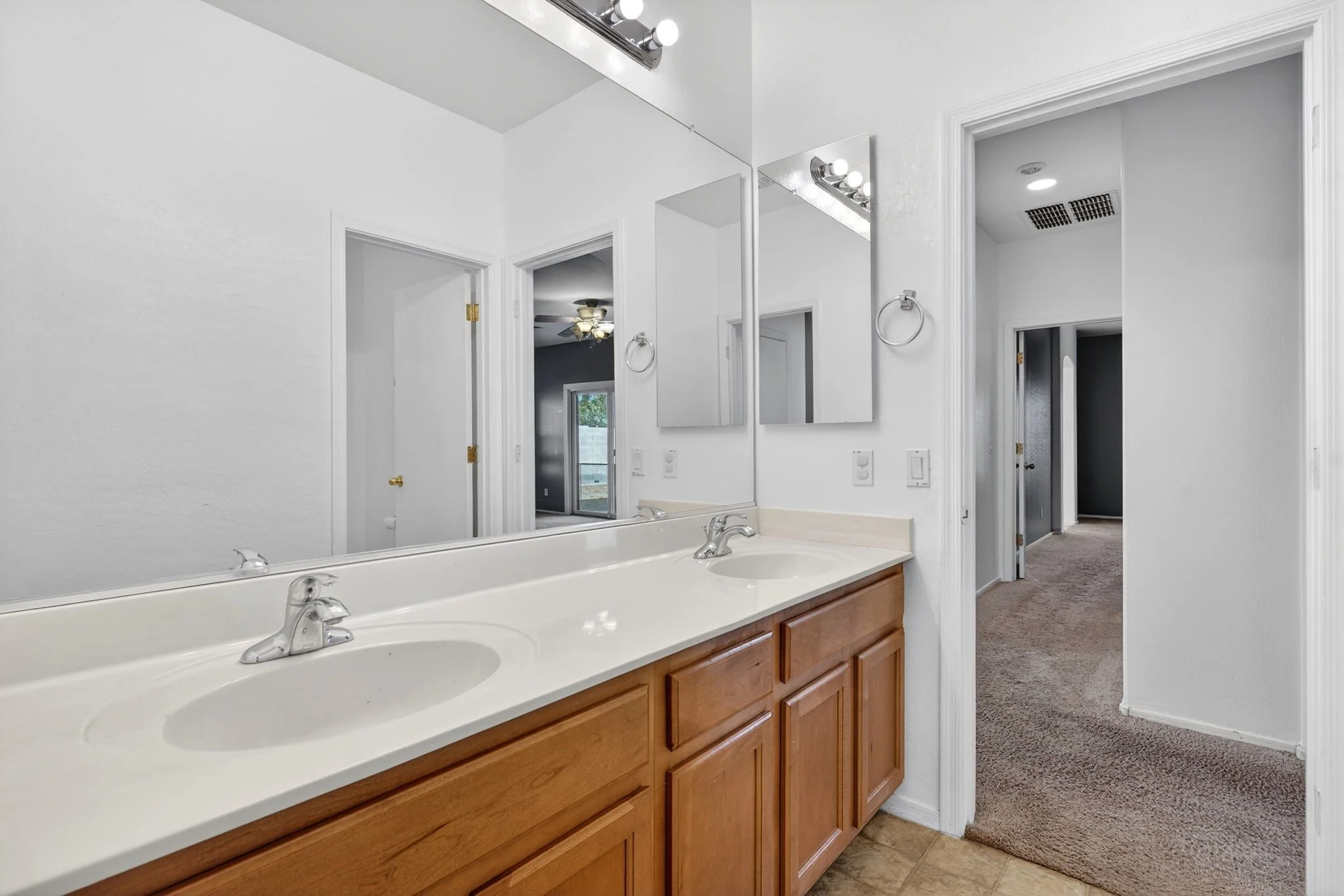 A bathroom vanity with double sinks, wooden cabinets, and a large mirror. The bathroom has white walls, recessed lighting, and a doorway leading to a carpeted room with a ceiling fan.