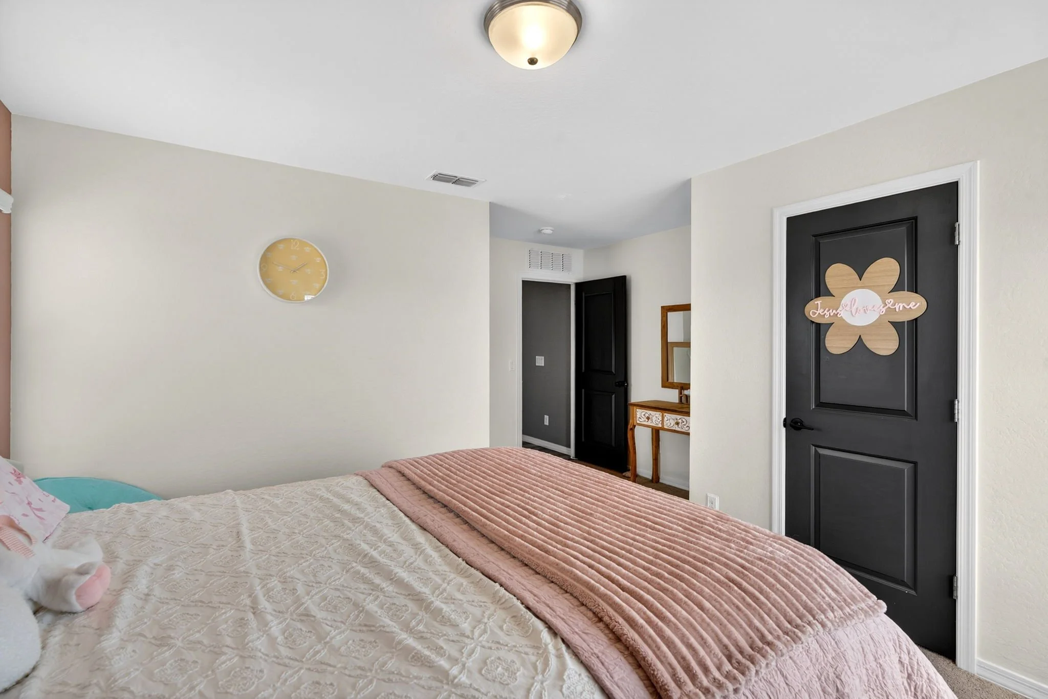 A bedroom with a bed covered in a white quilted blanket, pink throw and pillows, and a plush toy. There are off-white walls, a black door with a wooden butterfly-shaped sign reading 'Love, Dream, Me,' a wall clock showing approximately 10:12, a mirro