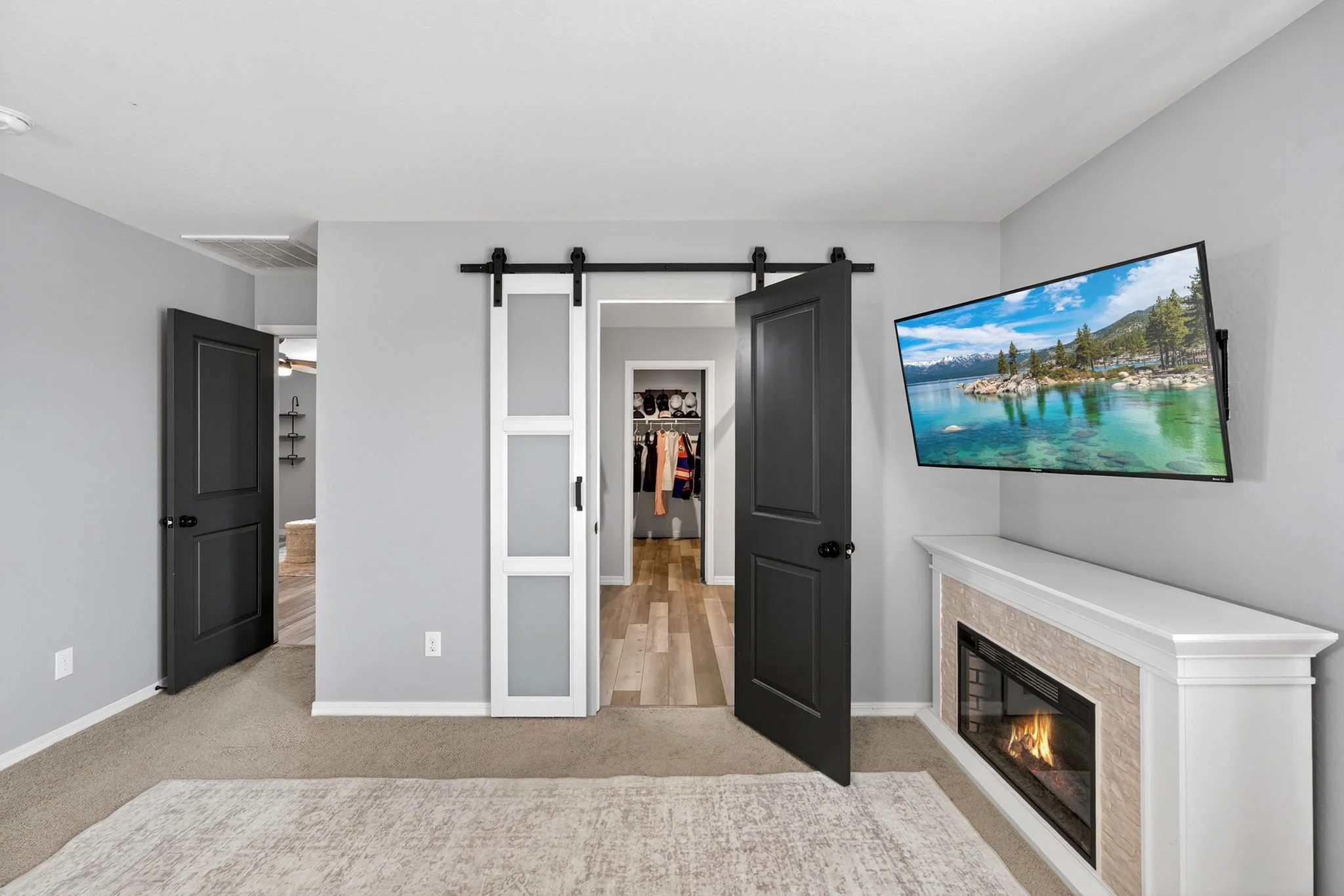 Interior of a modern living room with a wall-mounted TV displaying a scenic nature view, a fireplace with a lit fire, open dark gray doors, and a glimpse of a closet with clothes and shoes.