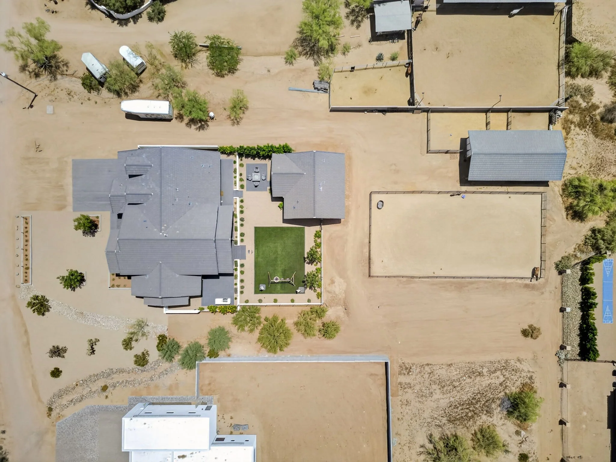 An aerial view of a residential backyard with a house, trees, a sandy yard, a small green patch, and a basketball court.