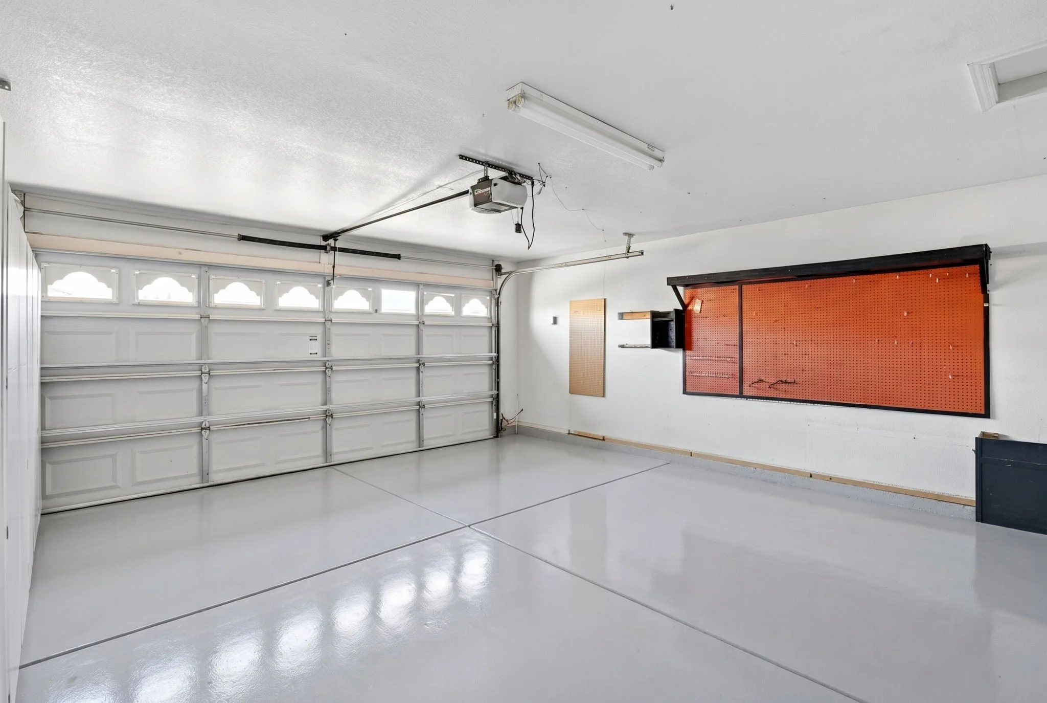 Interior of a clean, empty garage with a white roll-up door, overhead fluorescent lighting, and a pegboard on the wall.