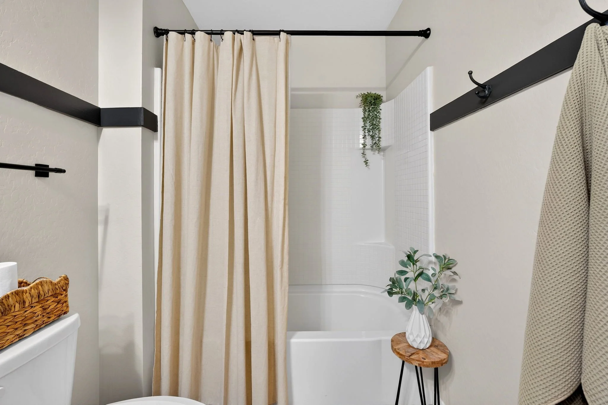 A bathroom shower with a cream-colored curtain on a black rod, white tiled walls, a small round wooden side table with a white geometric vase holding greenery, and a wicker basket on a white toilet.