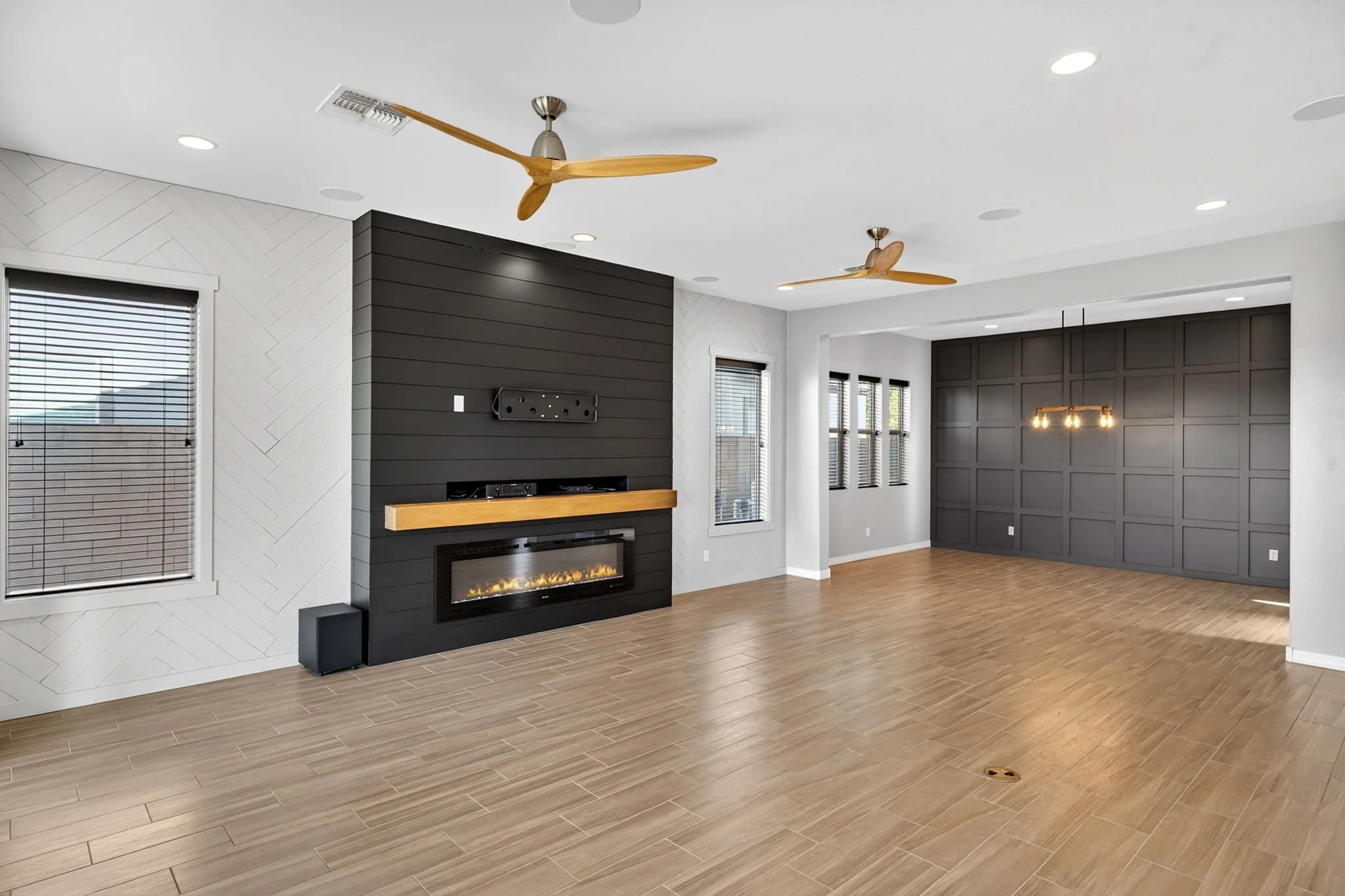 A modern living room with a white ceiling, light wood flooring, and large windows with blinds. There are two ceiling fans with wooden blades, a sleek black fireplace, and a gray paneled accent wall with a hanging light fixture.