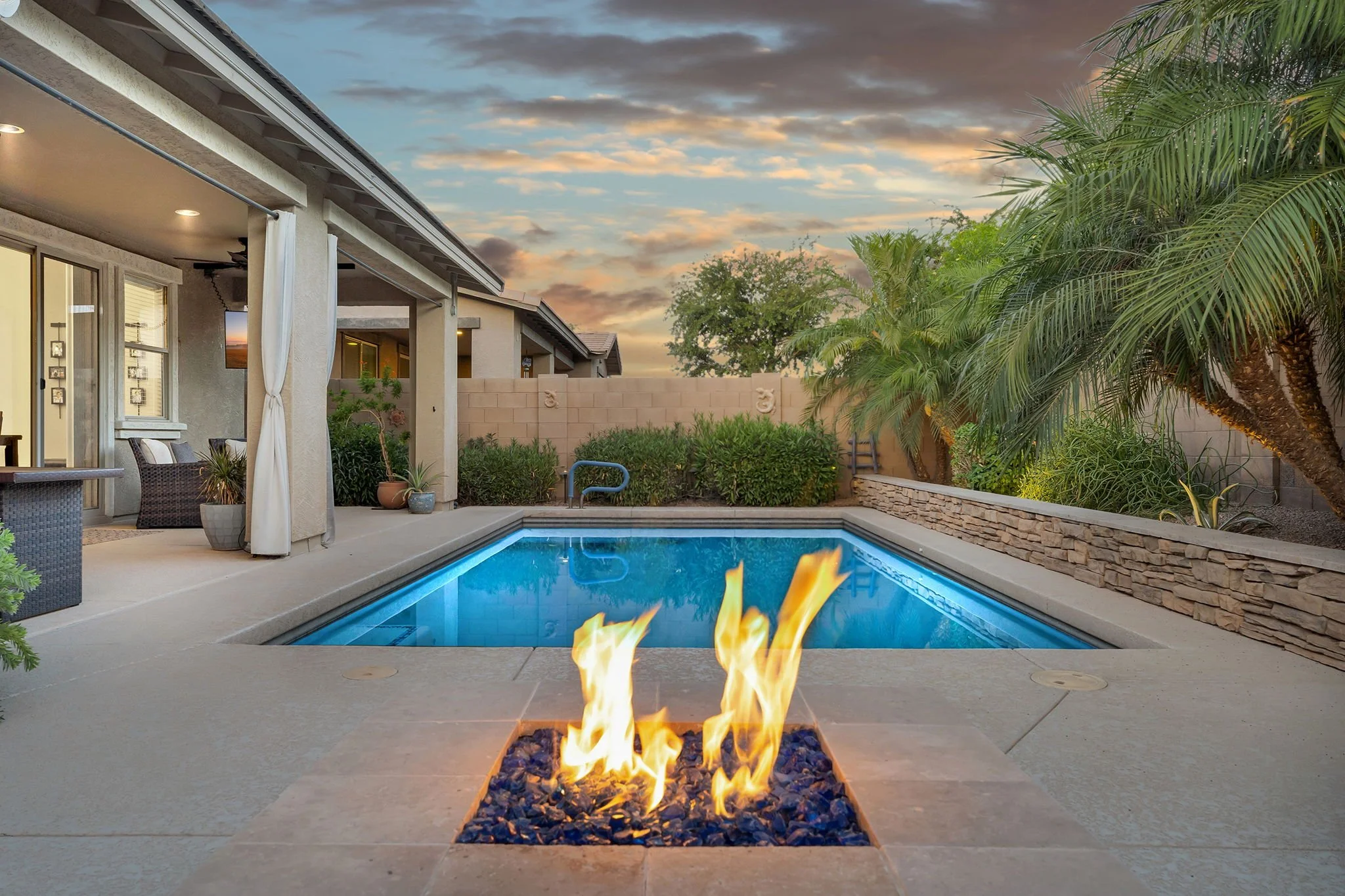 Backyard patio with a swimming pool, a fire feature, and surrounding greenery during sunset.