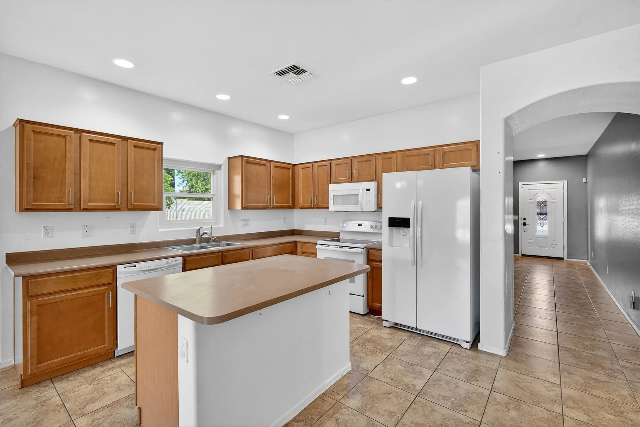 Modern kitchen with wooden cabinets, white appliances including a microwave and refrigerator, a double sink, and tiled flooring. There is a small island with a wooden top in the foreground and a hallway leading to the front door.