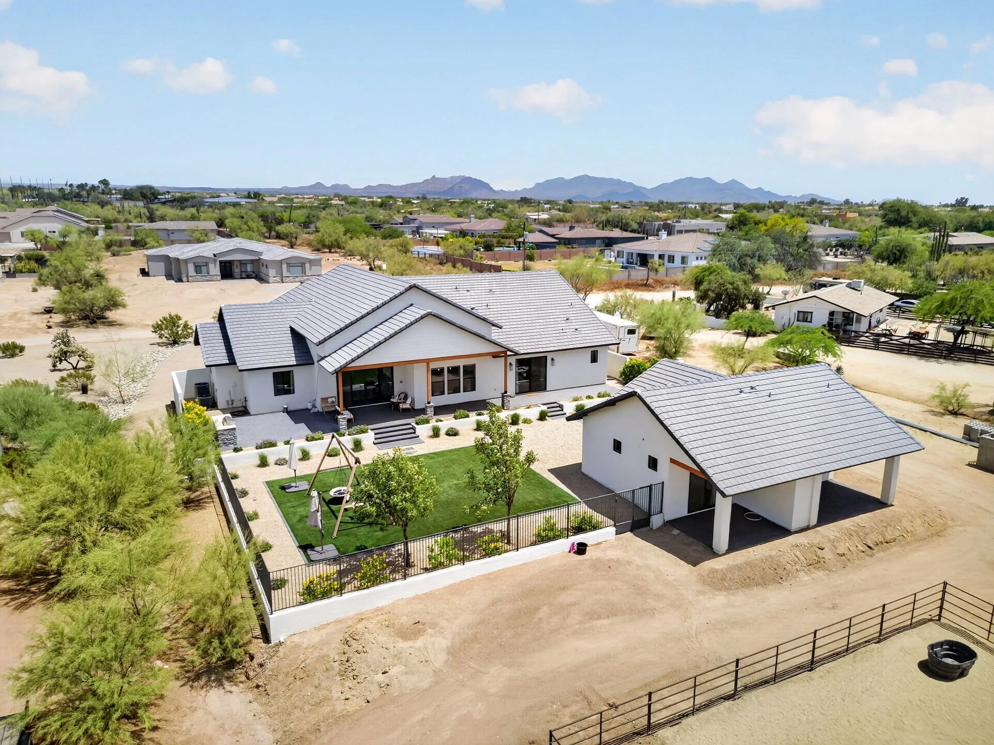 Aerial view of a residential backyard with a green lawn, trees, a swing set, and a white house, surrounded by a black fence, with neighboring houses and distant mountains in the background.