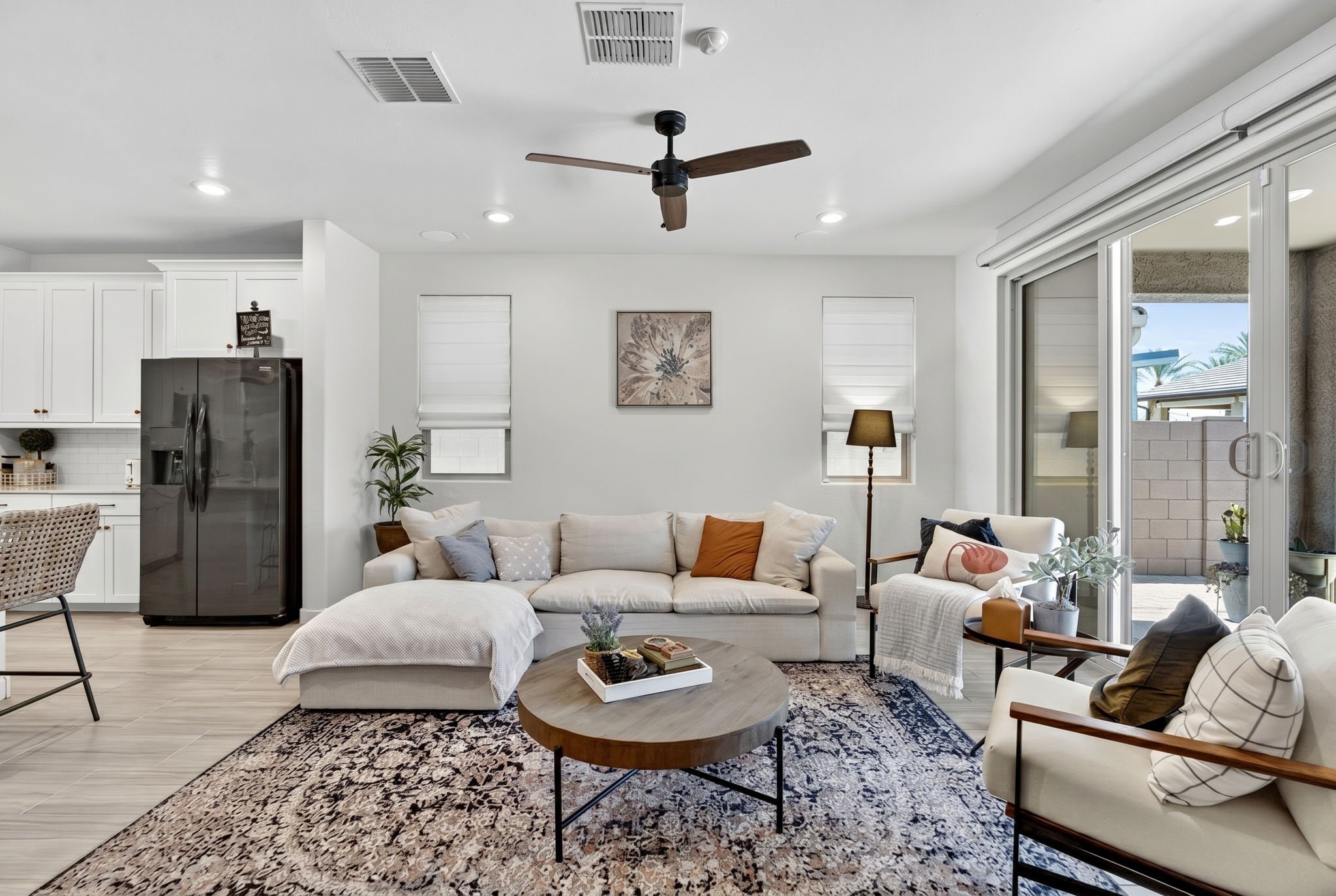 Living room with white sofa, armchair, coffee table, decorative pillows, area rug, potted plants, artwork, ceiling fan, large sliding glass door, and kitchen in the background.