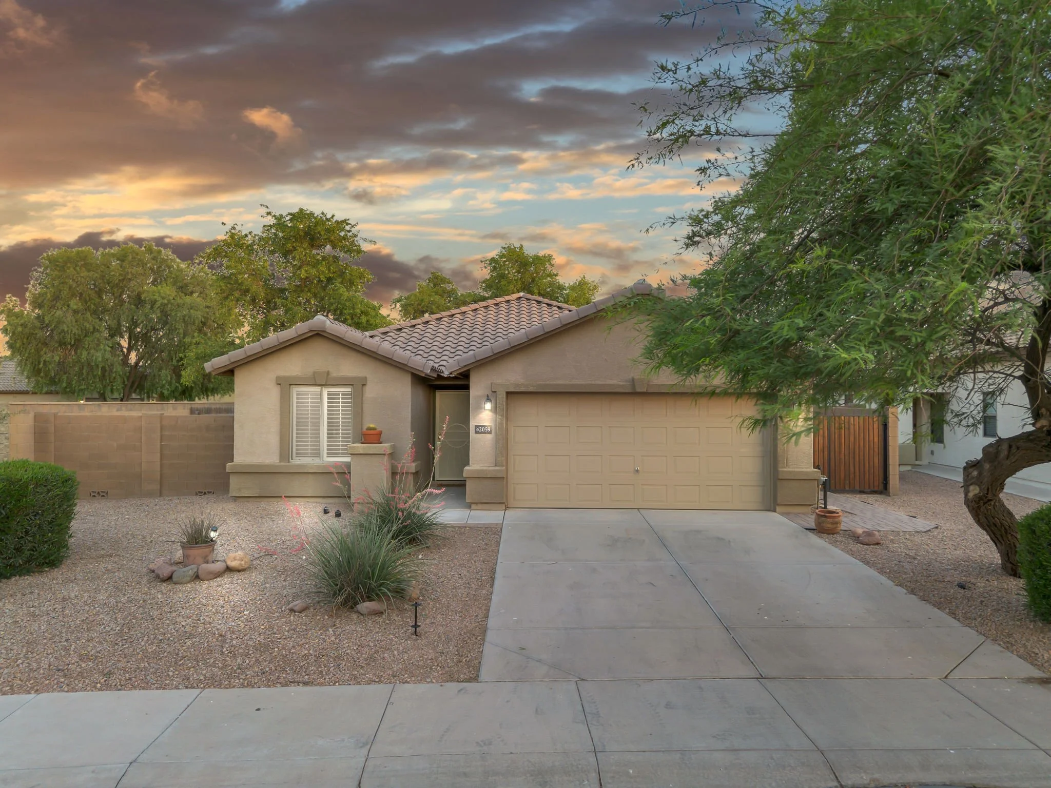 A suburban house with a tiled roof, a closed garage door, a front entrance with a small porch, and desert landscaping in the yard. The sky is partly cloudy at sunset.