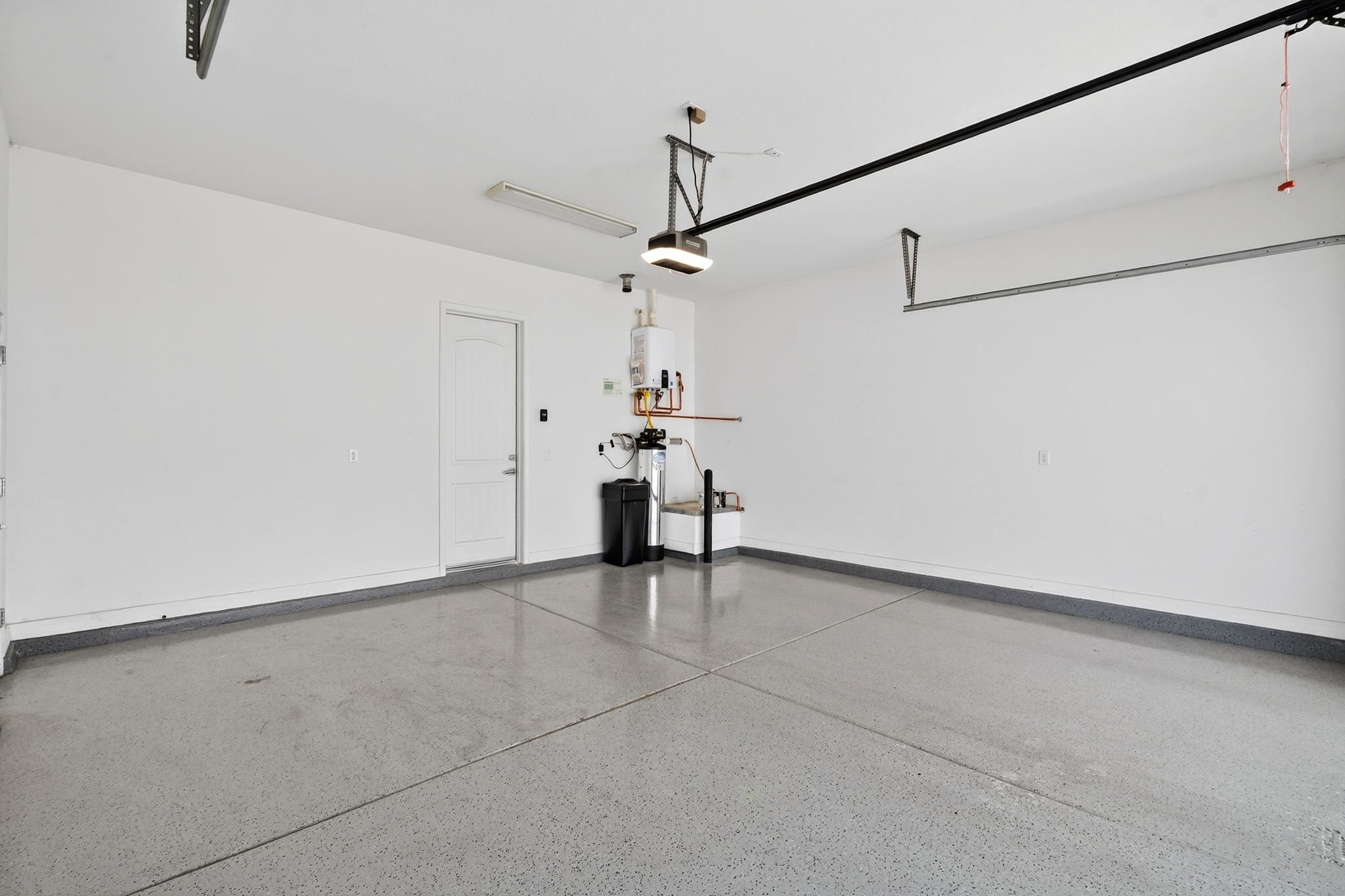 Empty garage with white walls, grey concrete floors, overhead lights, garage door tracks, and some wall-mounted utility equipment.