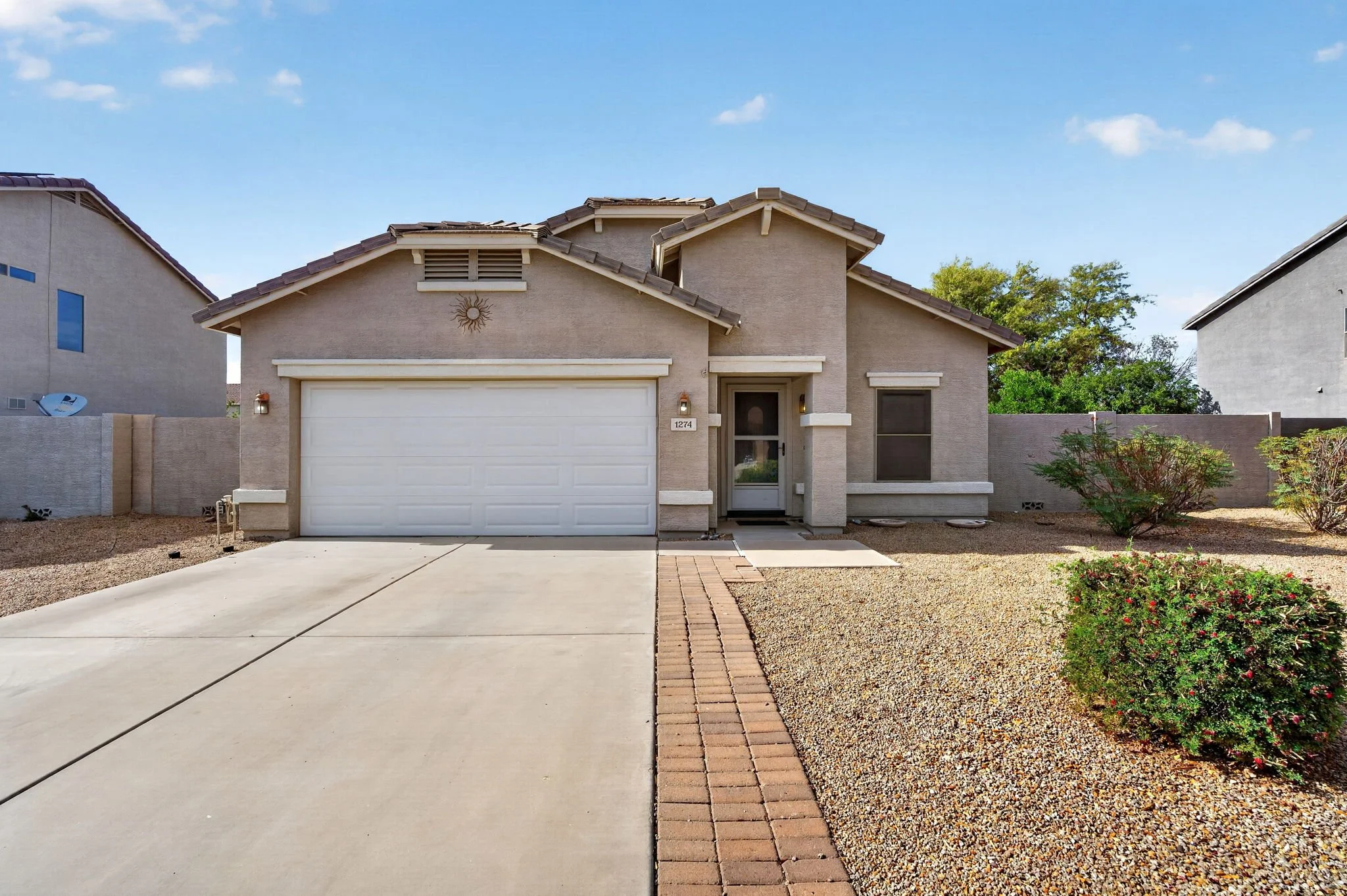 A modern suburban house with a two-car garage, beige stucco exterior, and a small front yard containing gravel and some plants. The house has a front door, small windows, and a decorative sun sculpture above the garage. The sky is blue with a few clo