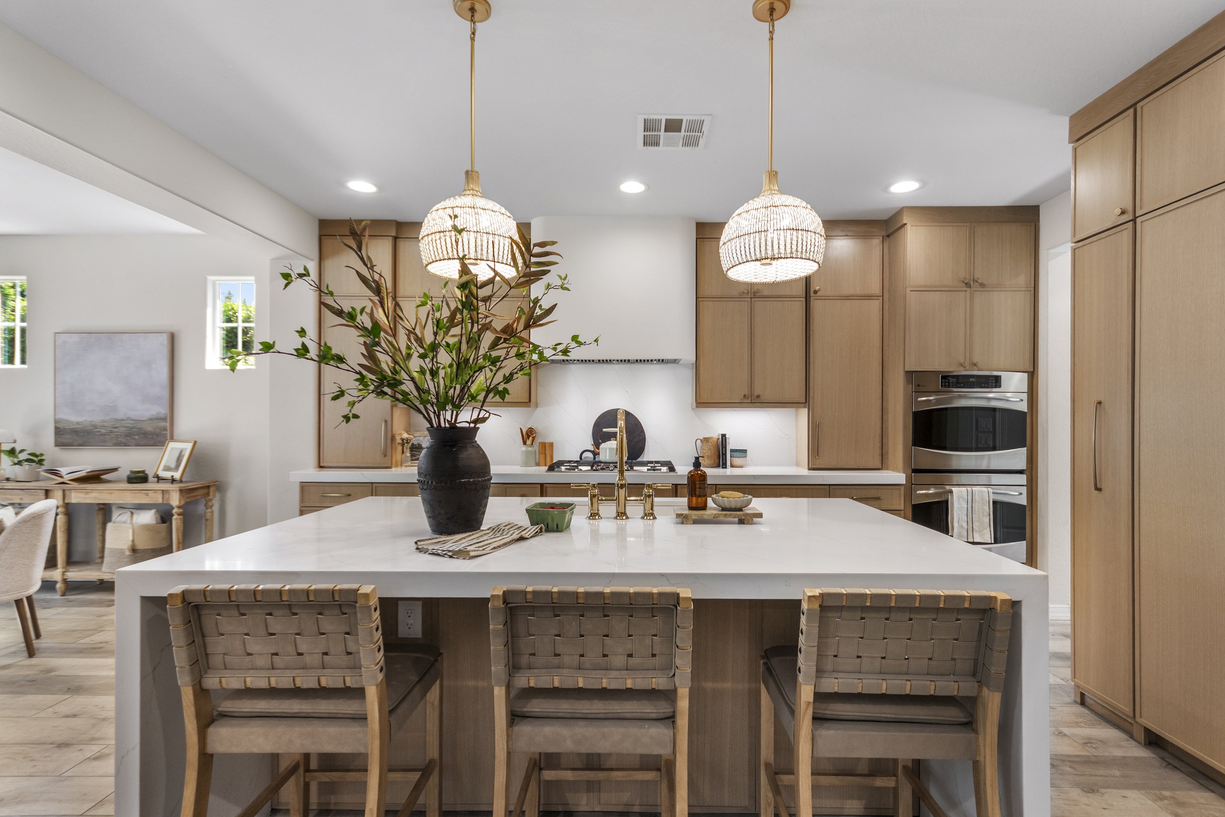 Modern kitchen with white island, wooden cabinetry, pendant lights, decorative plants, and built-in oven.