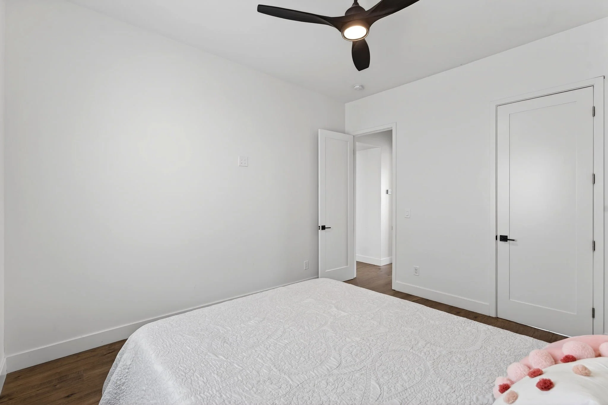 A bedroom with white walls, a ceiling fan with light, a partially visible bed with a white quilt, hardwood floors, and closed white doors.
