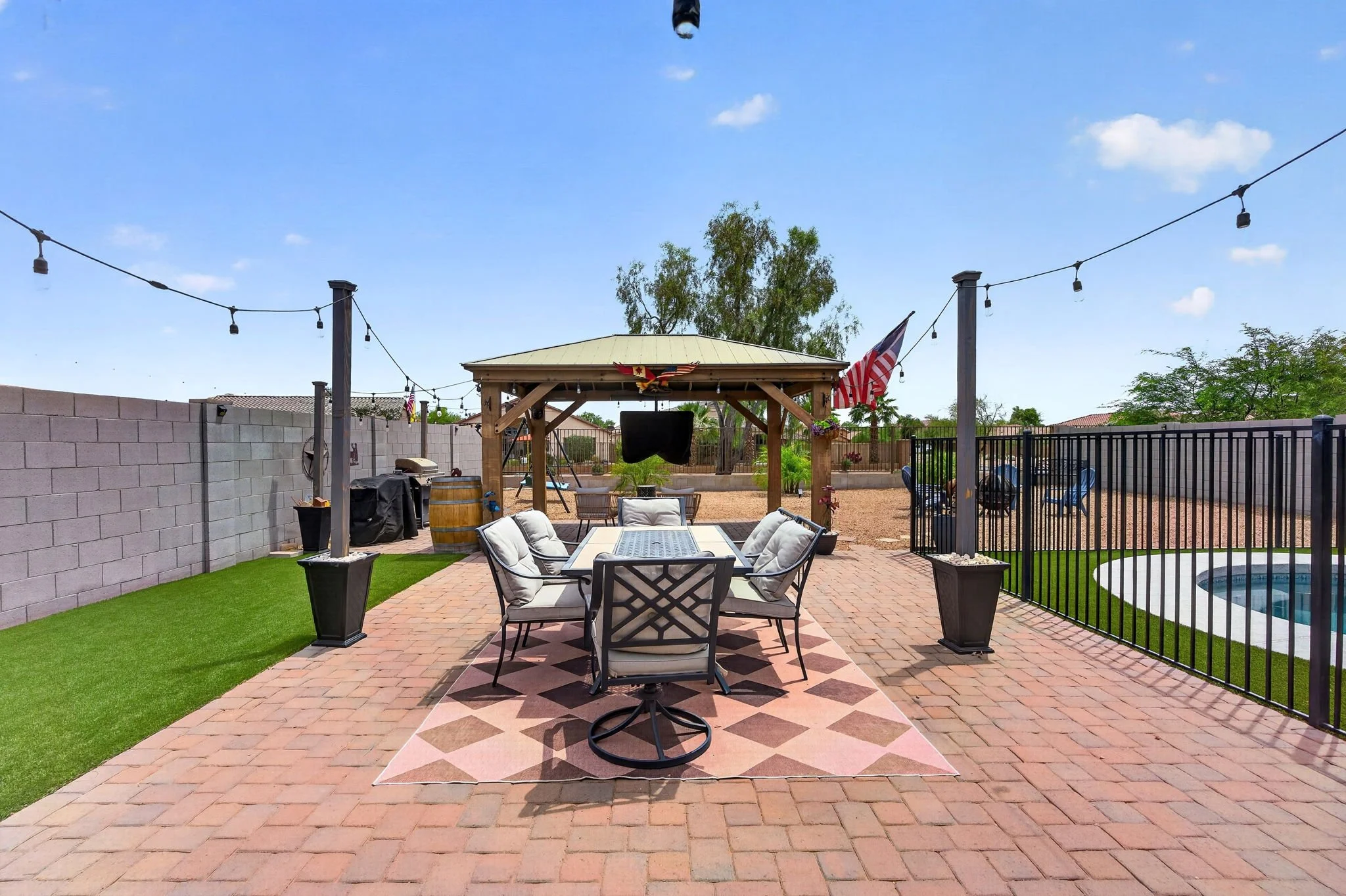 Backyard patio with outdoor dining table and chairs, a gazebo with a flag and a bird sculpture, string lights, artificial grass, a concrete block wall, and a fence enclosing a small pool.