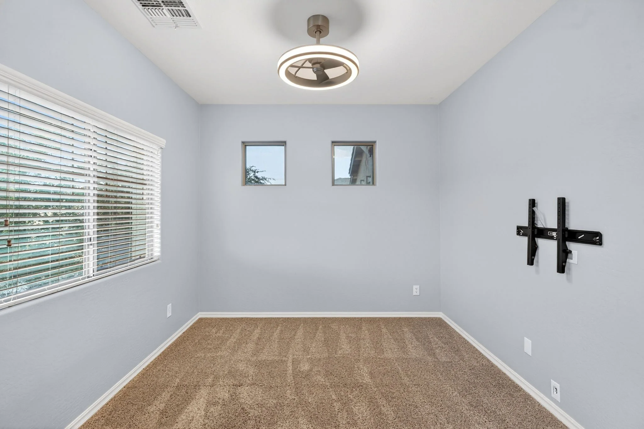 Empty room with beige carpet, light blue walls, three small windows on the back wall, a large window with white blinds on the left, and a mounted TV bracket on the right wall. Ceiling fan with circular light fixture.