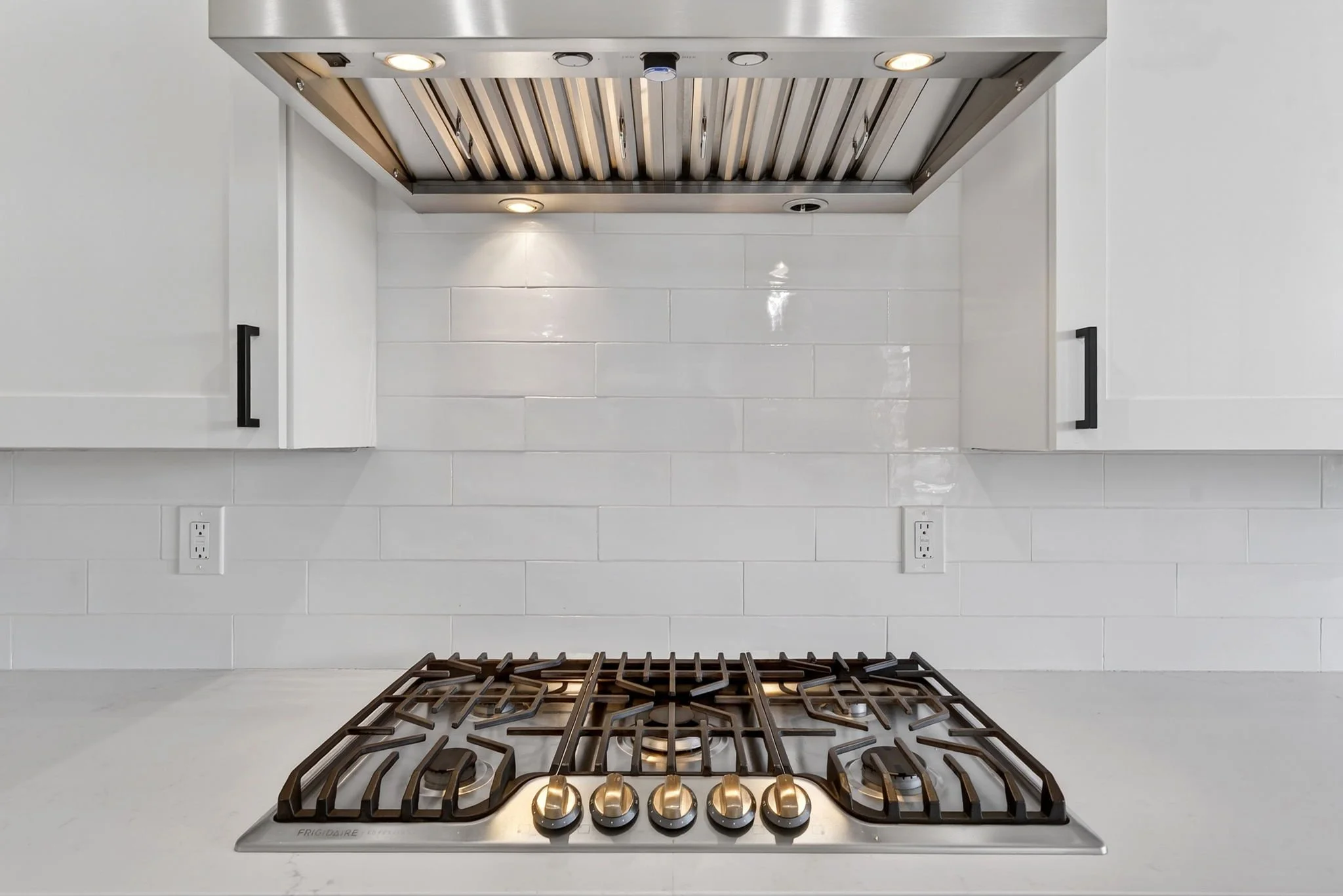 A modern kitchen stove with a gas cooktop featuring four burners, set against a white tiled backsplash and white cabinets with black handles.