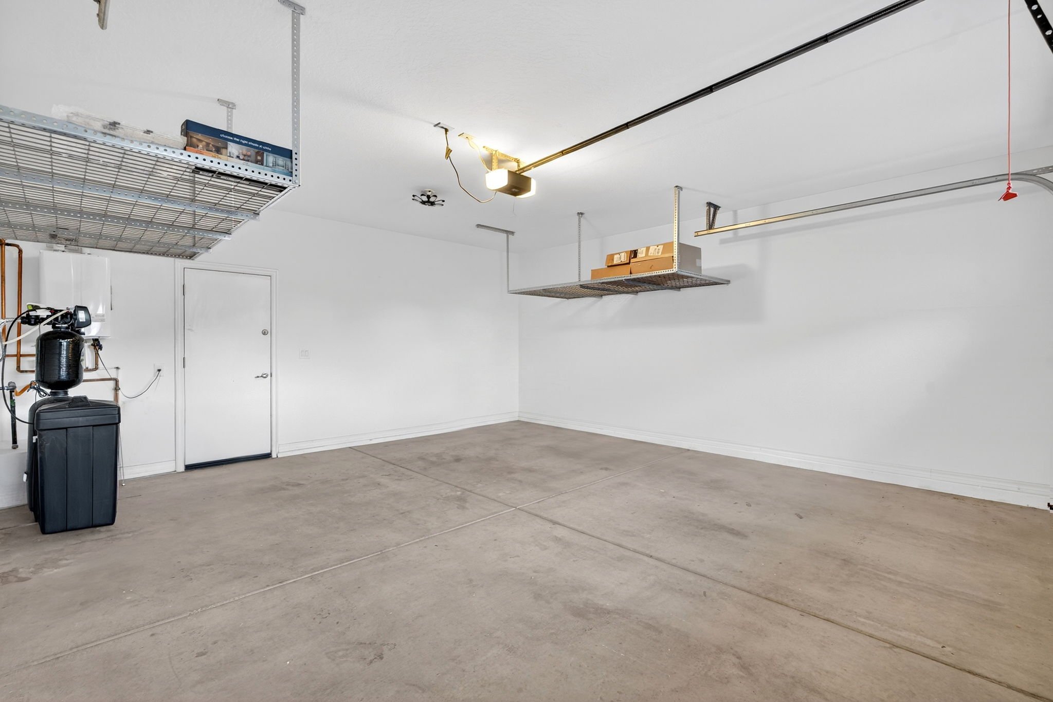 Empty garage with a concrete floor, white walls, and a white ceiling. There are ceiling-mounted resistances, a metal shelf with boxes, and a water softener system on the left side.