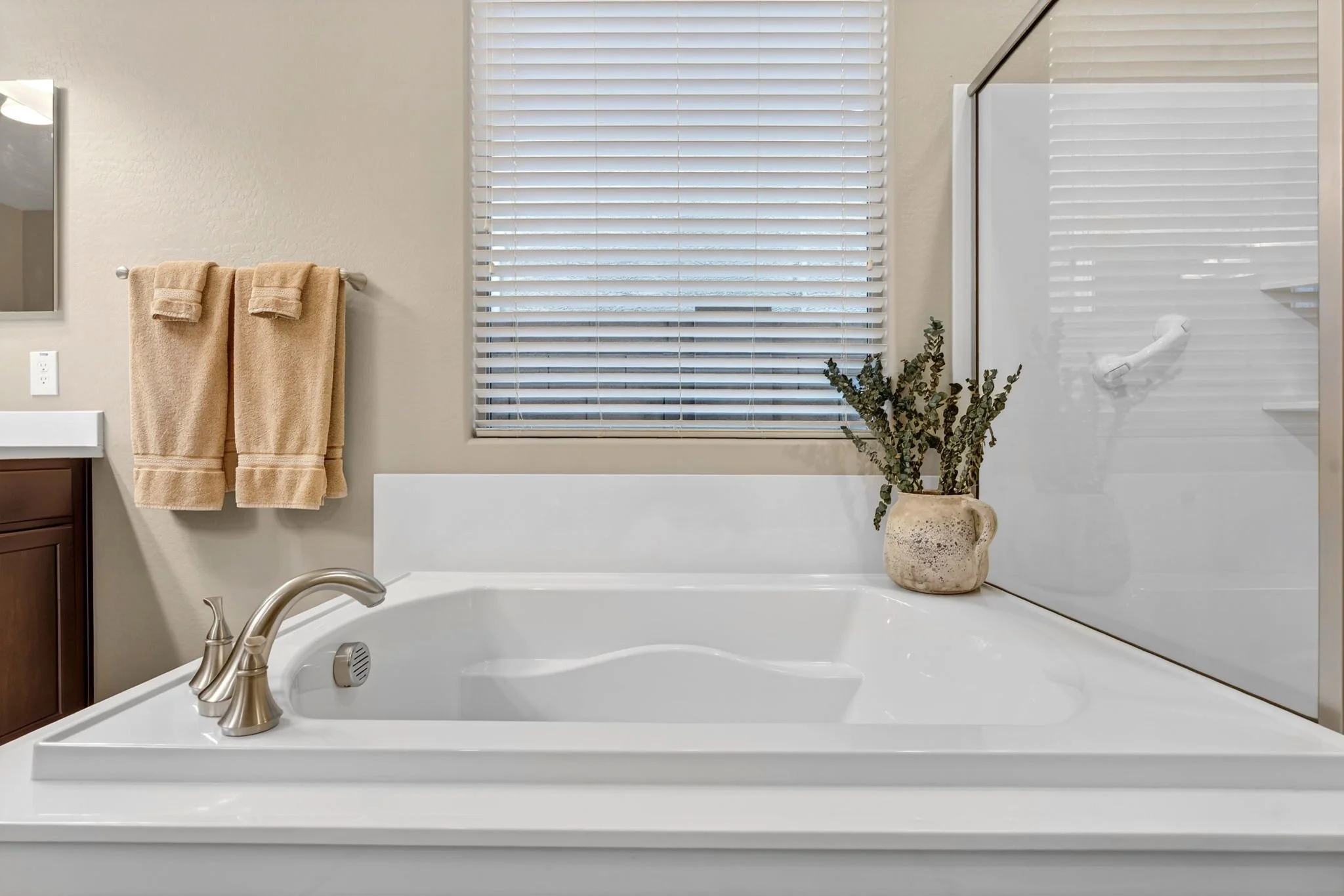 Bathroom with a bathtub, faucet, a potted plant in a beige vase, a window with white blinds, beige towels hanging on a towel bar, and a mirror on the left side.