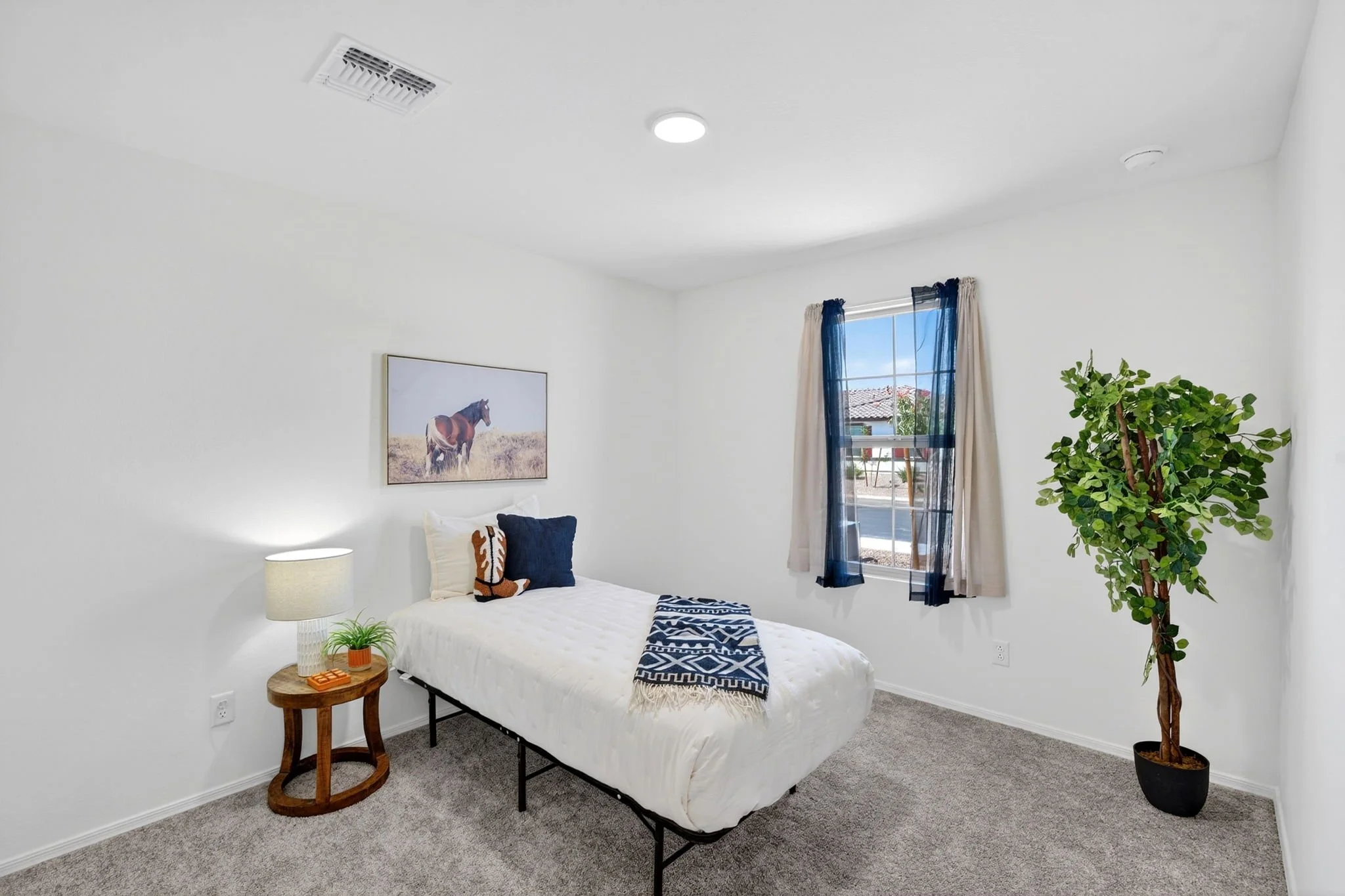 A bedroom with white walls, a bed with white bedding and pillows, a wooden side table with a potted plant and a remote, a window with beige and navy curtains, and a tall green houseplant.