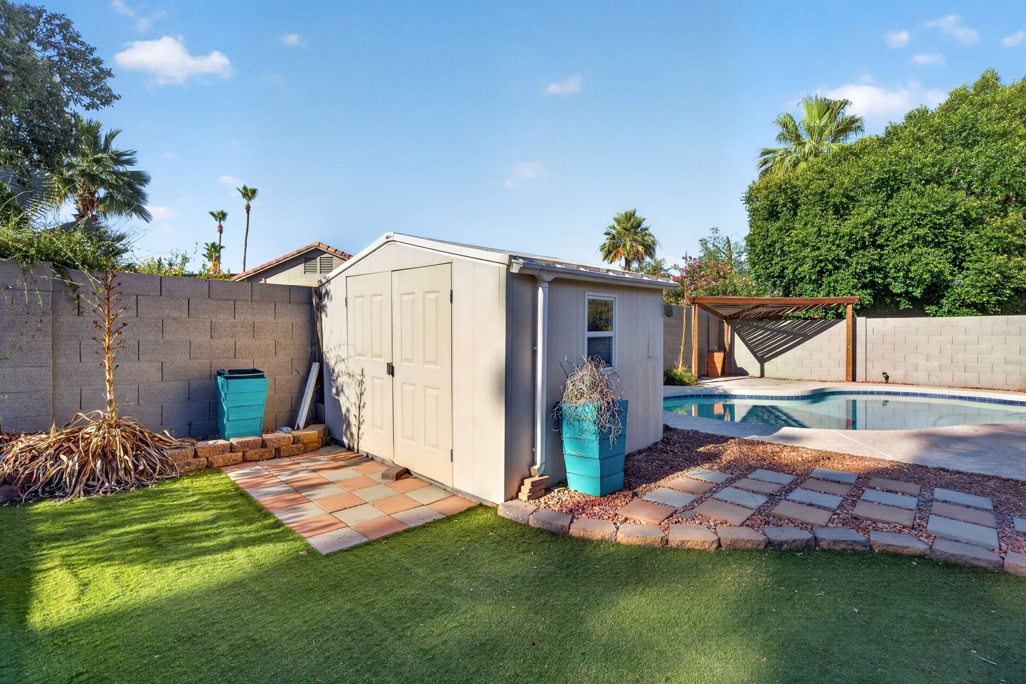 Backyard with artificial grass, a concrete block wall, a small shed, a swimming pool, and landscaping with plants and trees under a clear blue sky.