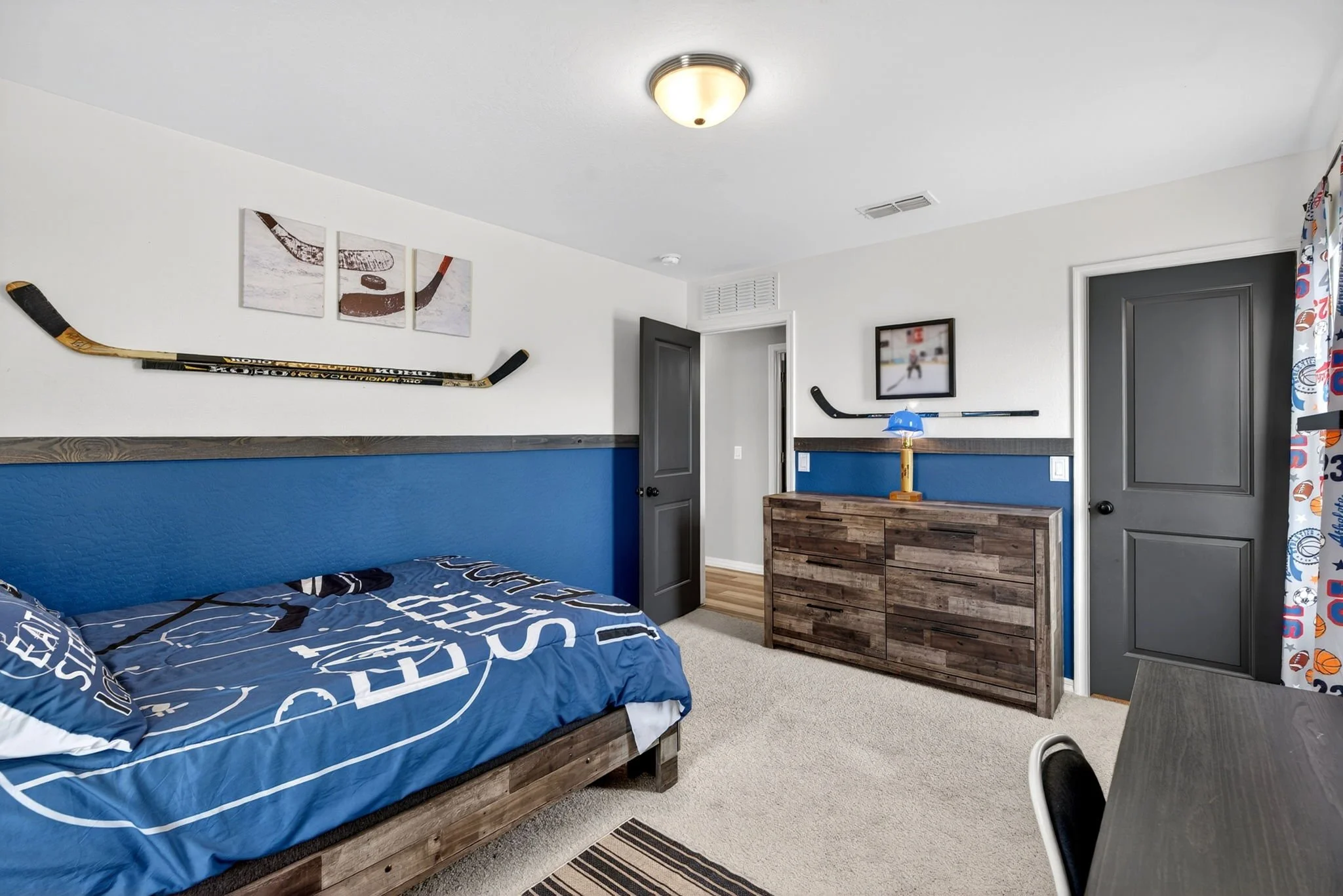 A bedroom with white walls, a gray door, and hockey sticks mounted on the wall. There is a bed with blue hockey-themed bedding, a wooden dresser with a blue table lamp, and a framed hockey game picture on the wall. Curtains with sports patterns are p