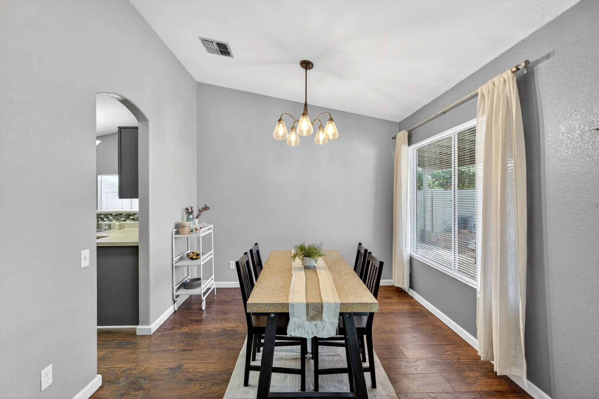 A dining room with a long rectangular table, six black chairs, a small white rolling cart, a window with white curtains, a chandelier with six glass shades, and hardwood floors.