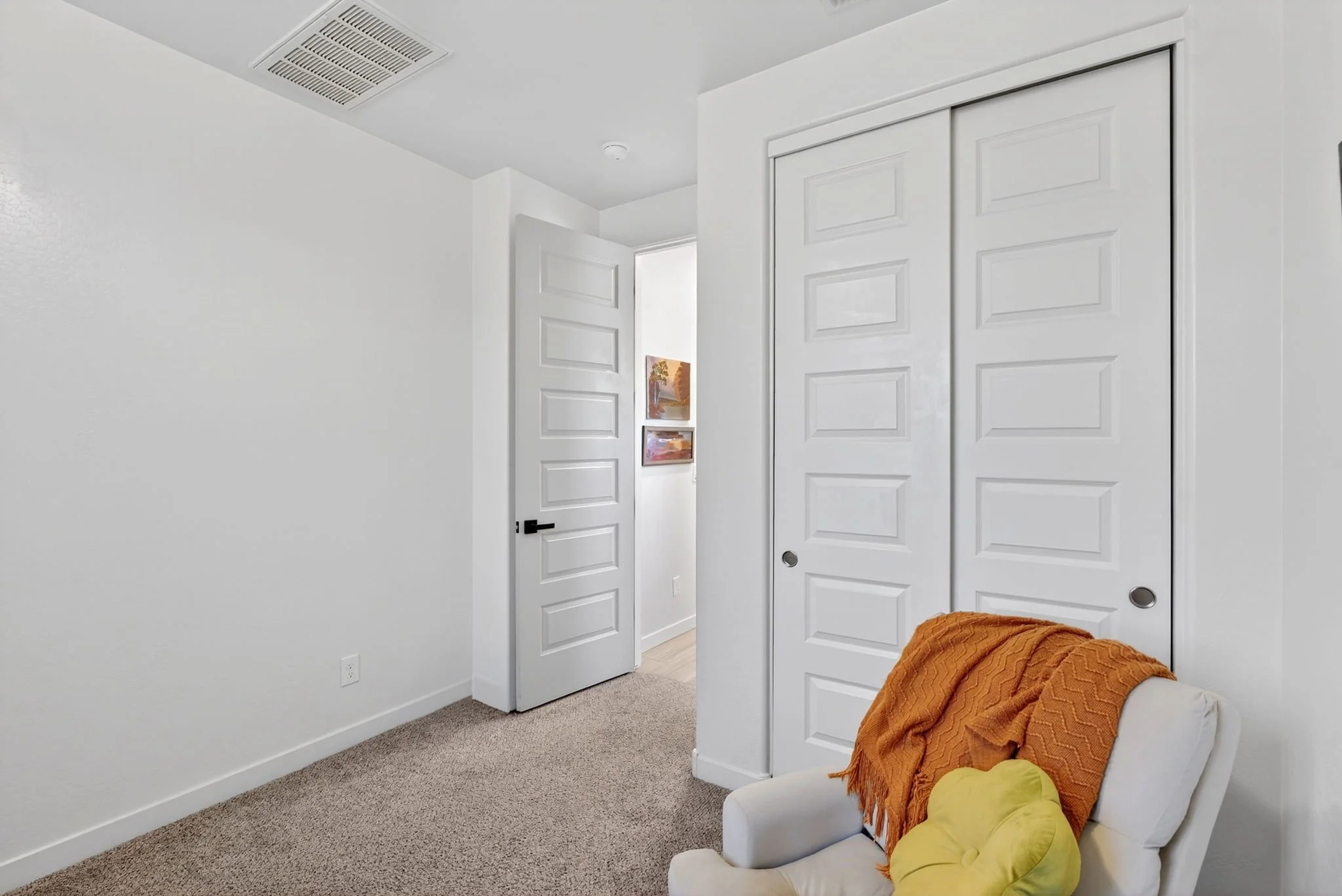 A bedroom with white walls, a closet with sliding doors, a white door slightly open, a small painting on the wall, and a bed with an orange blanket and a yellow pillow.