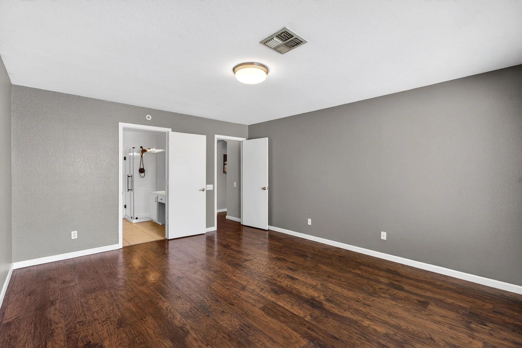 Empty room with hardwood floors, gray walls, ceiling light, and open doors leading to other rooms.