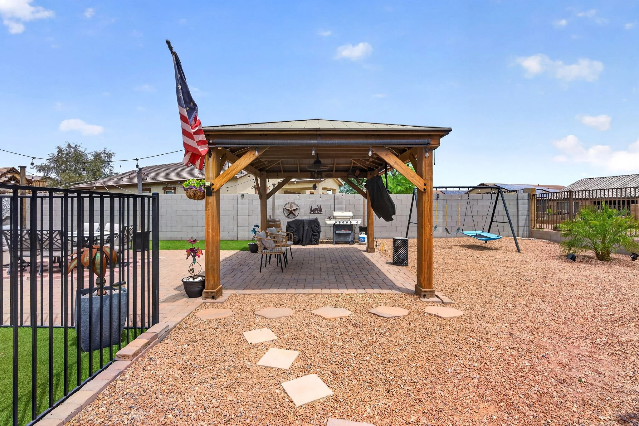 Backyard patio area with a wooden pergola, outdoor furniture, a barbecue grill, a children's swing, decorative plants, and string lights under a blue sky.