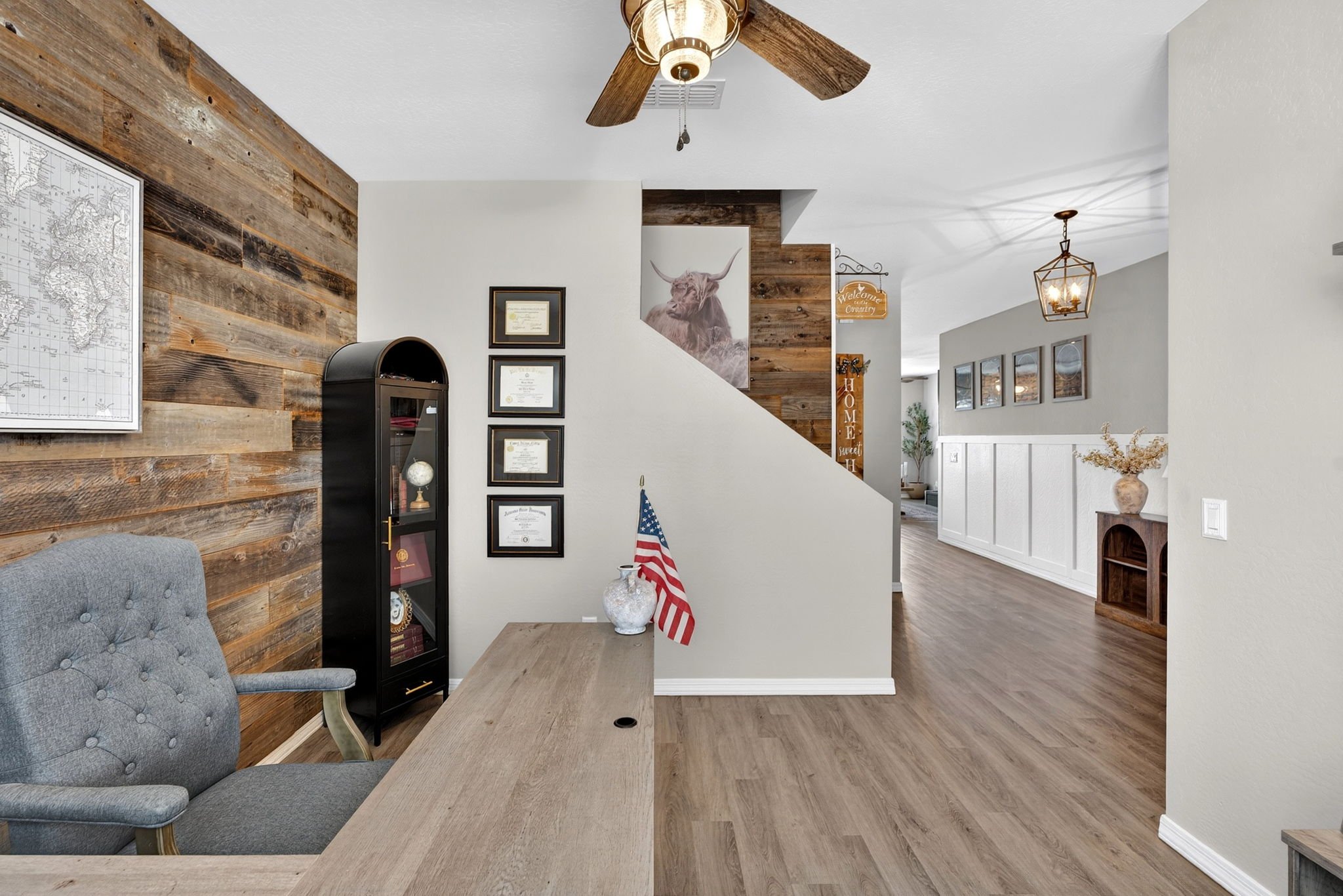 Interior of a home with wood-paneled walls, a ceiling fan, framed certificates, a decorative bull painting, and discounts. There is a desk with an American flag, and the area extends to a dining space with a hanging lantern light and framed pictures 